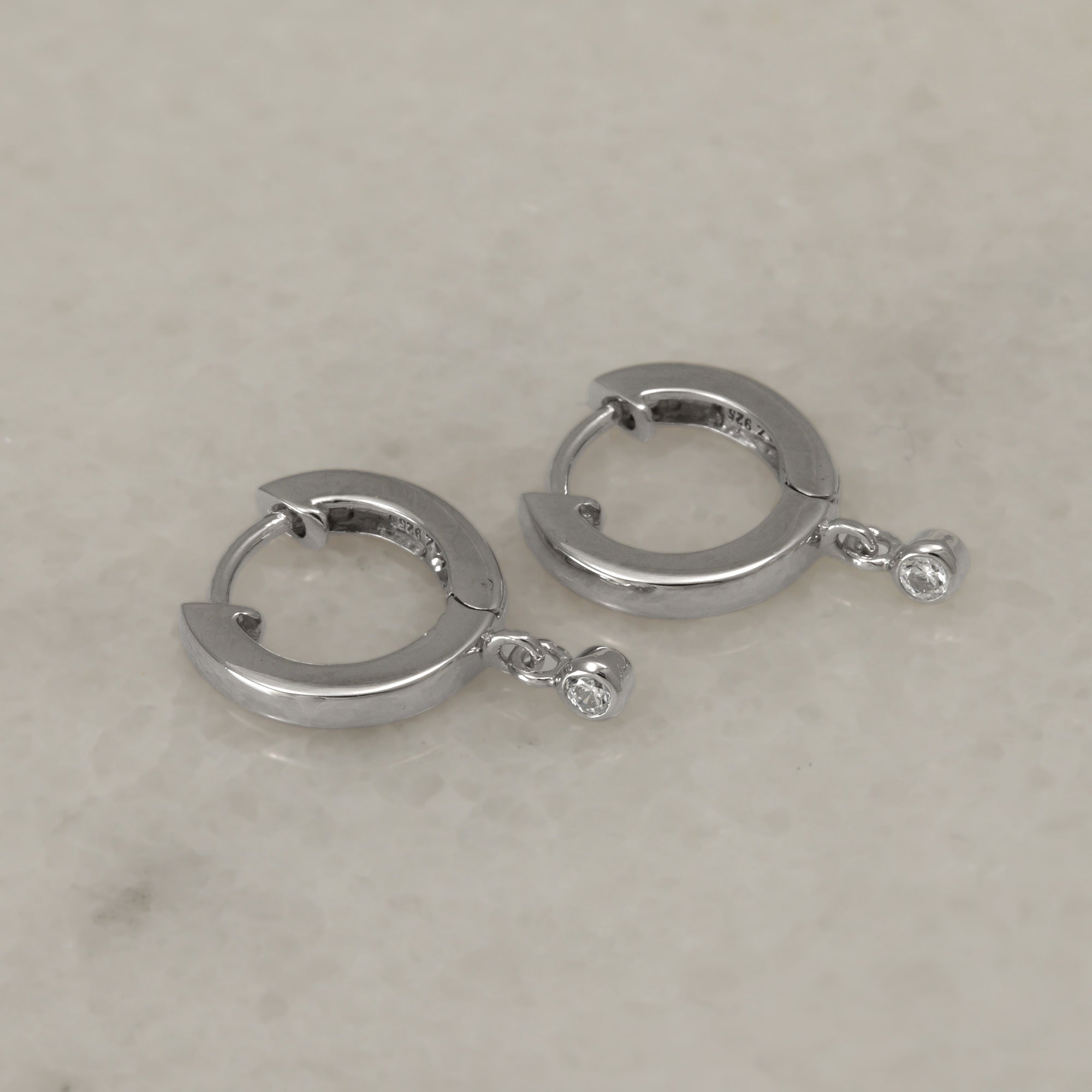 Sterling silver hinged hoop earrings featuring a delicate hanging sparkling stone, combining minimalist design with refined elegance in contemporary silver jewellery by Azendi.