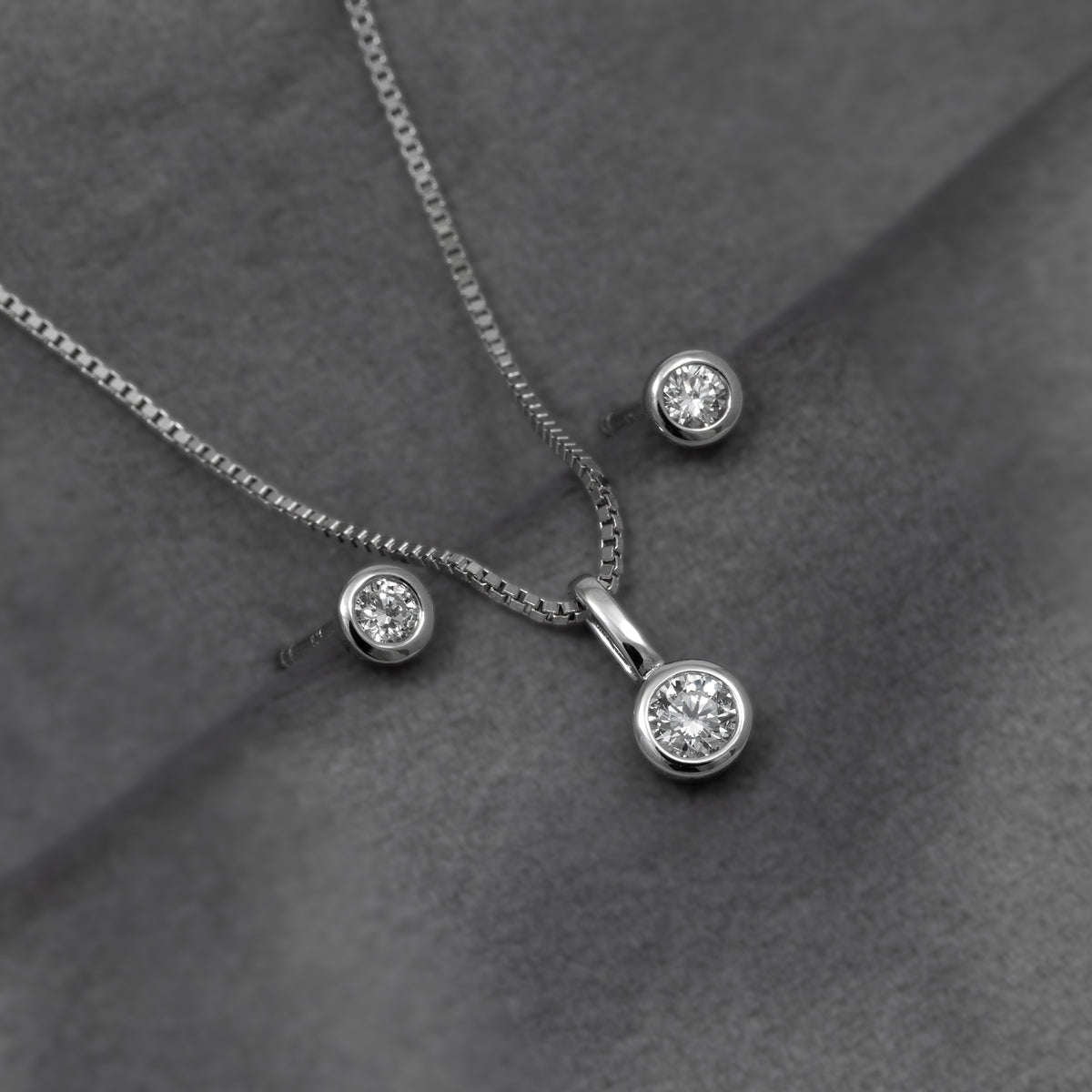Silver necklace with a round bezel-set diamond pendant, shown with matching stud earrings on a dark gray textured background.