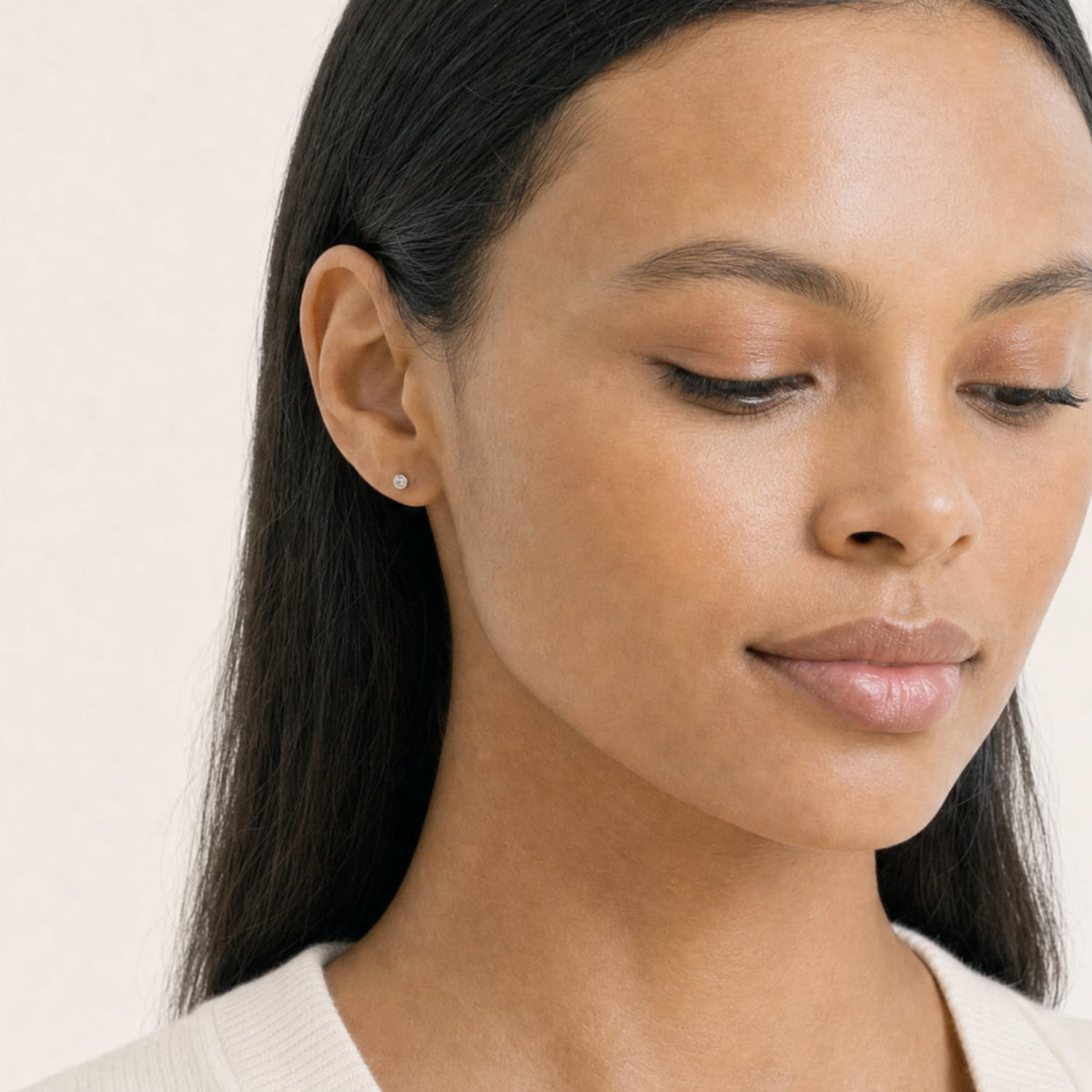 A close-up of a woman with dark hair wearing a delicate silver and lab grown diamond stud earring, set against a soft, light beige background.