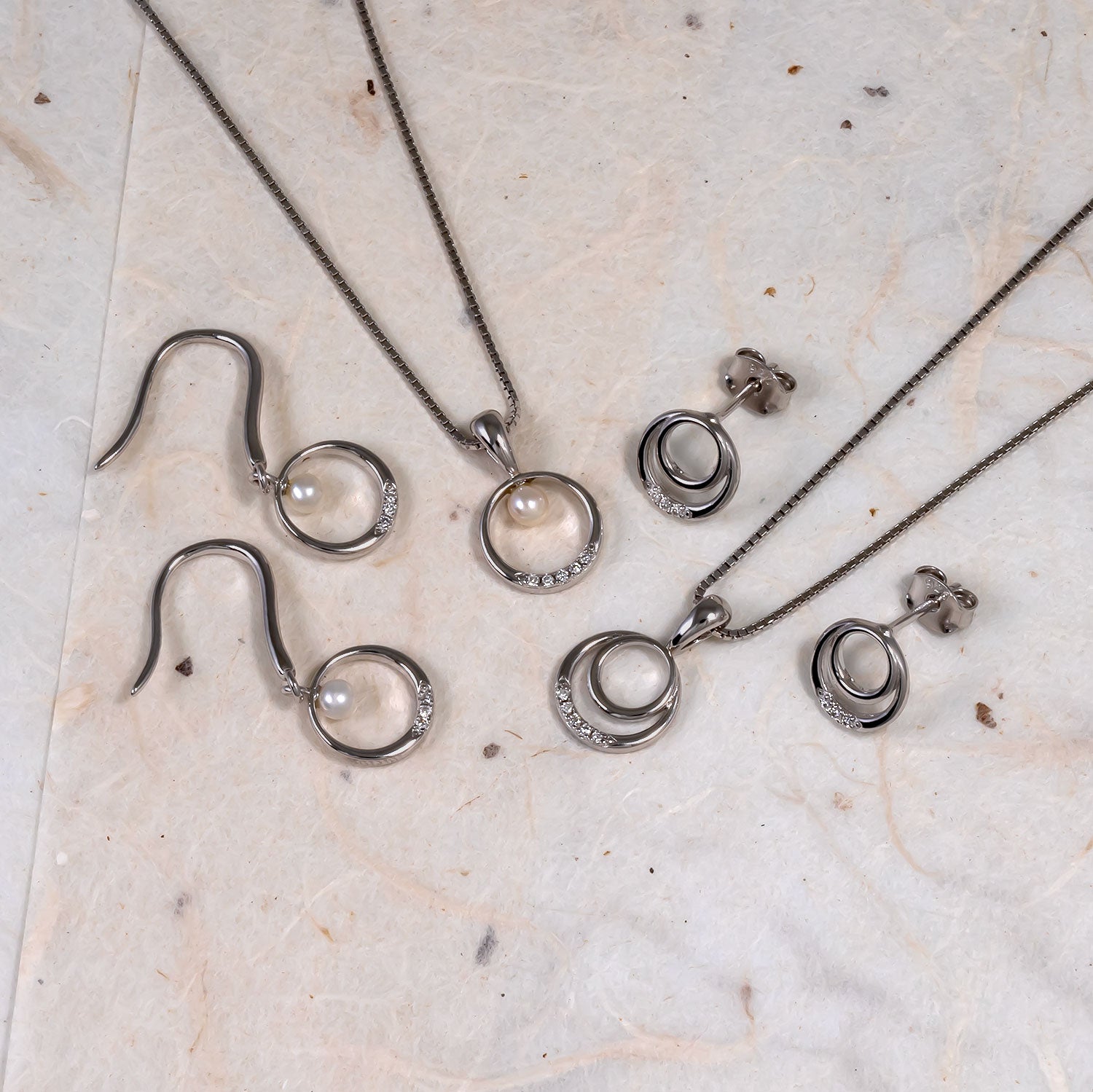 Collection of sterling silver jewellery including two pendant necklaces and matching pairs of earrings featuring sculptural double circle designs accented with small lab-grown diamonds on a light marble surface.