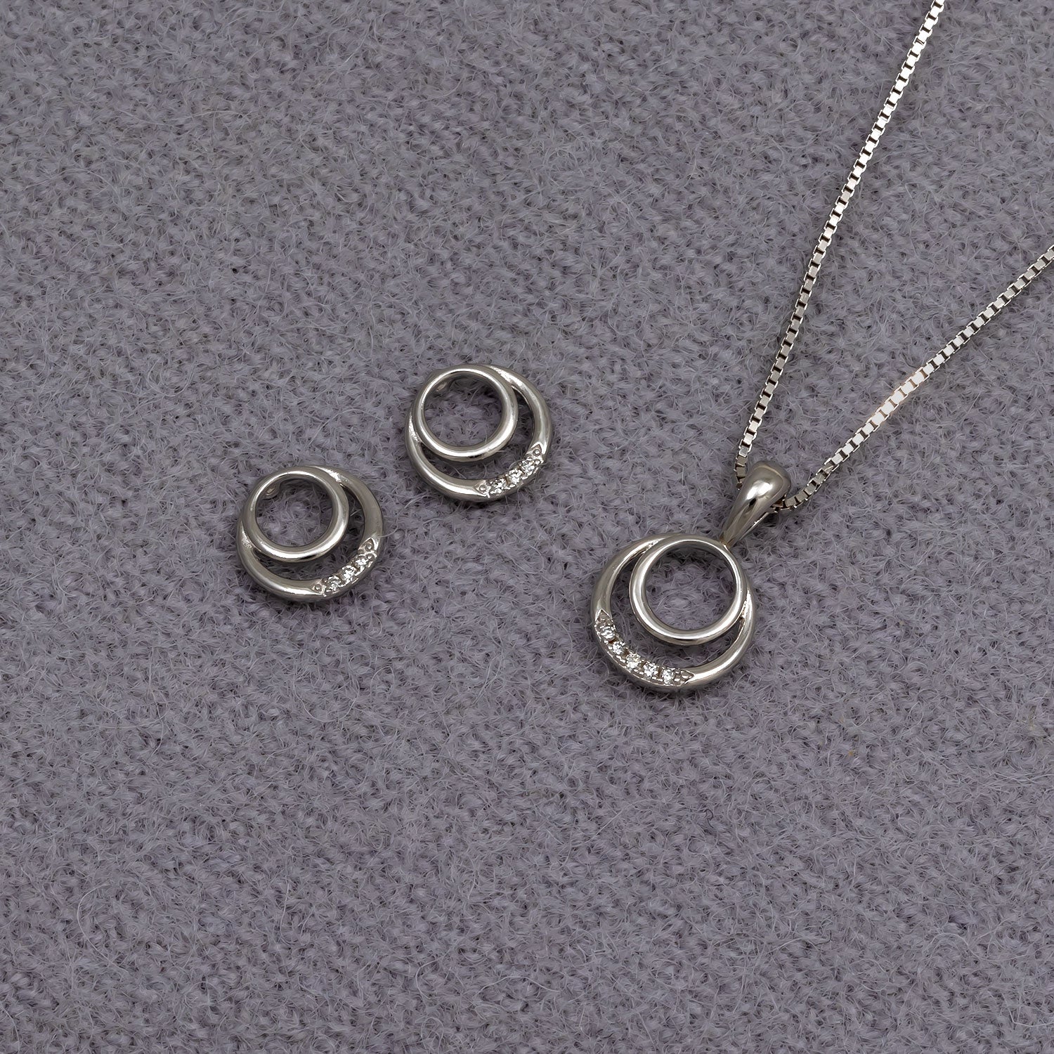 Sterling silver stud earrings and matching pendant featuring intertwined circles accented with small lab grown diamonds, presented on a soft grey fabric background.