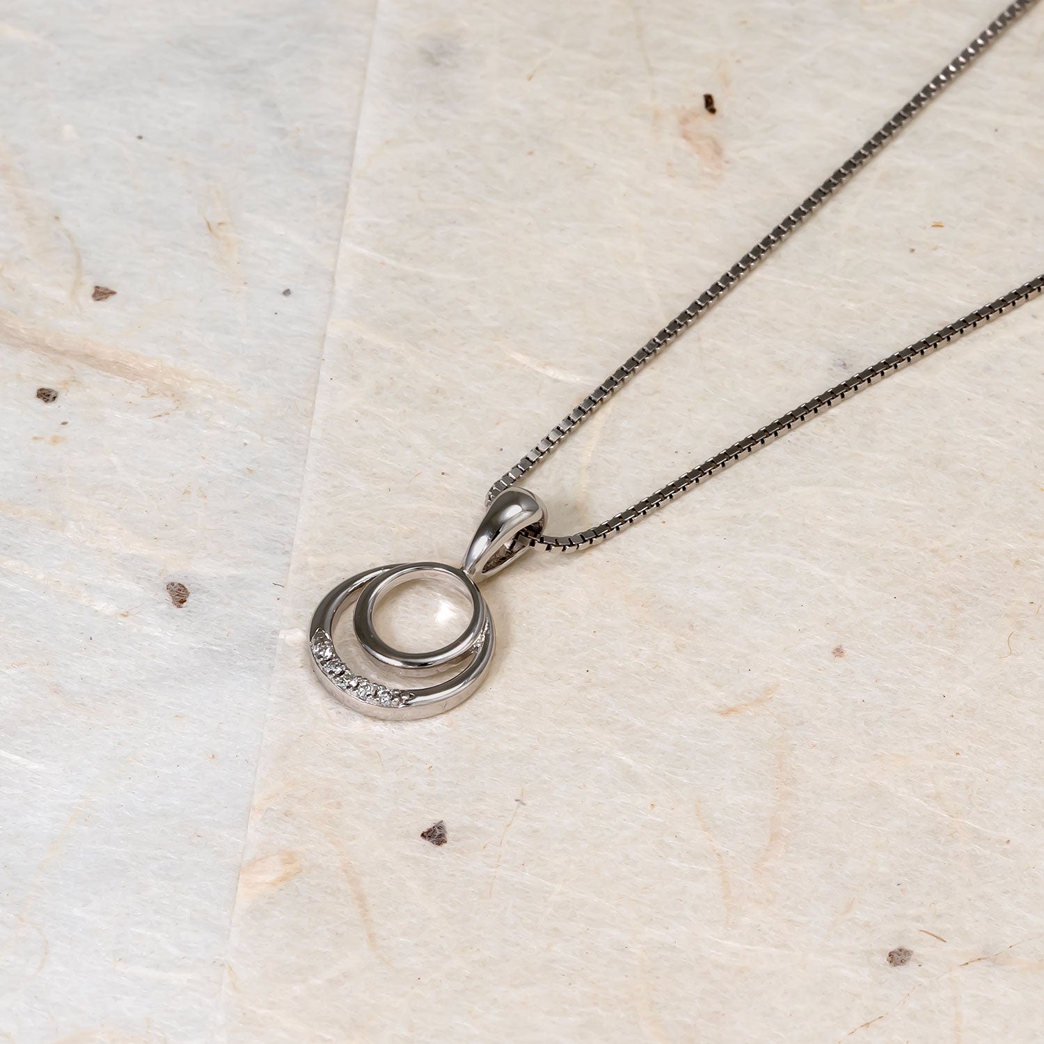 Sterling silver pendant featuring two interlocking circles, one adorned with lab grown diamonds, suspended from a fine box chain on a natural textured surface.