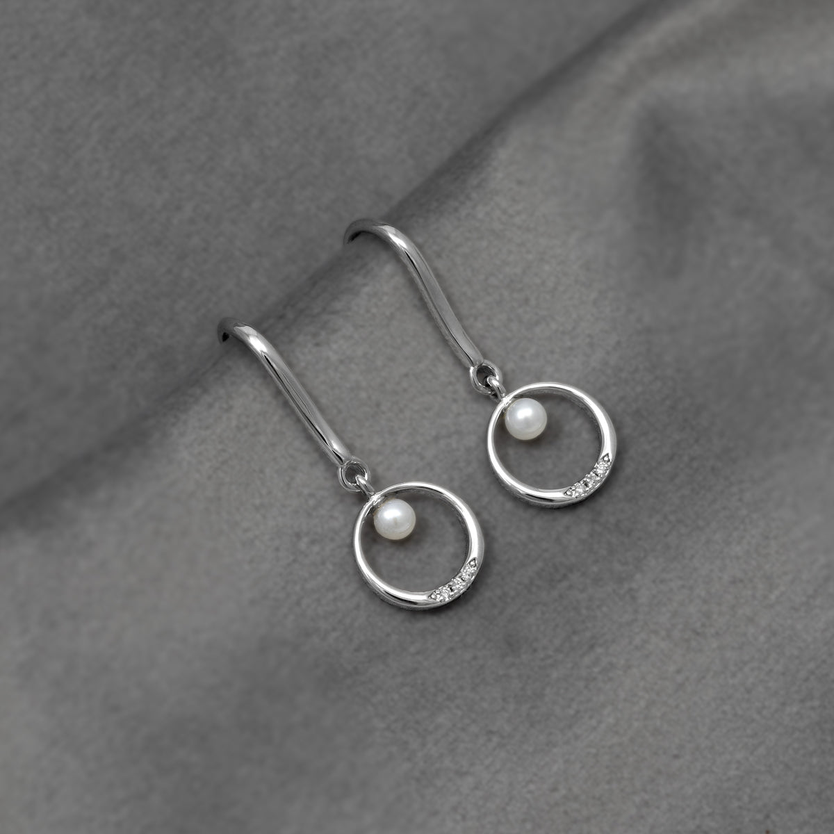 Elegant sterling silver drop earrings featuring small freshwater pearls and delicate lab-grown diamond accents within smooth circular frames, displayed on soft textured fabric.