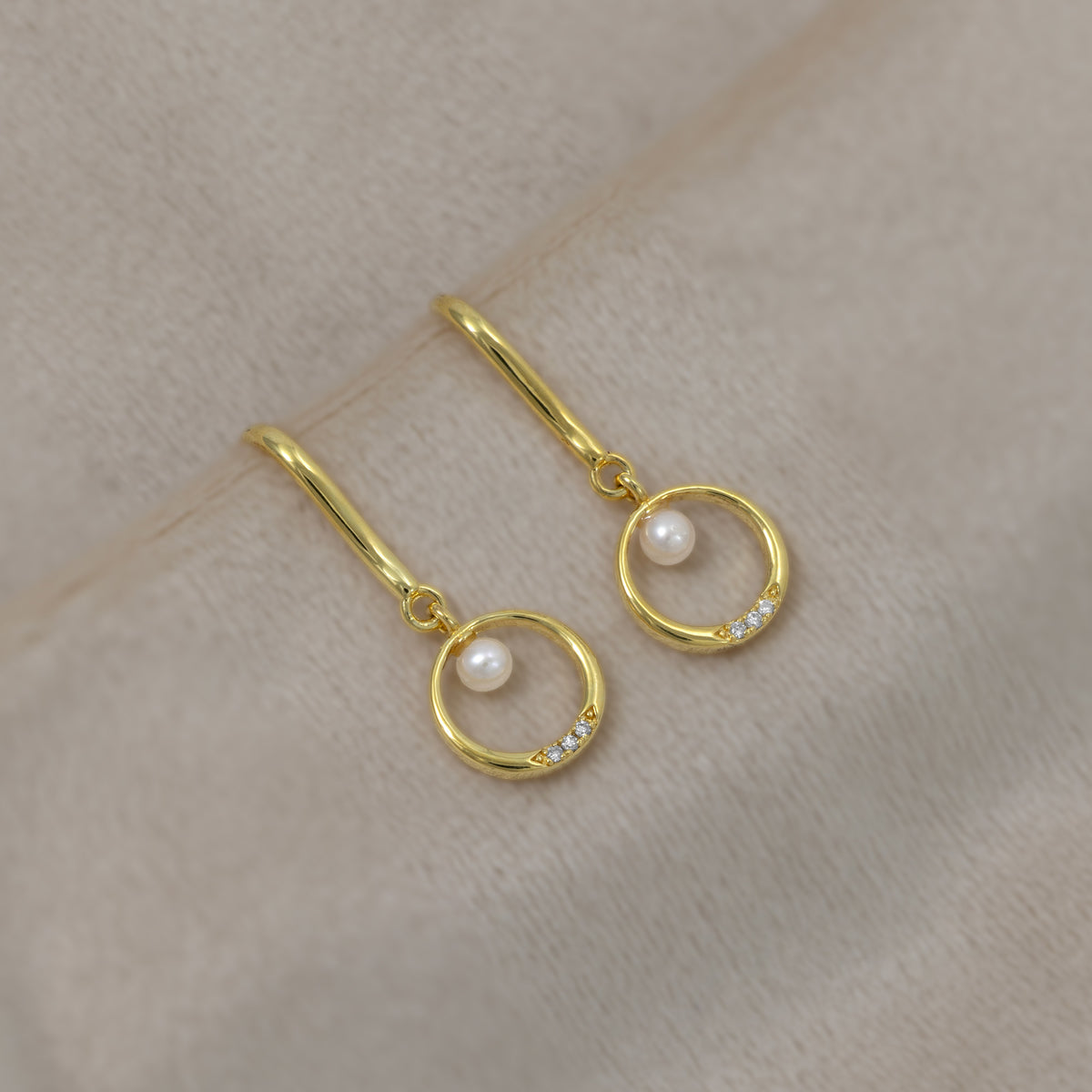 Elegant gold vermeil drop earrings featuring a small freshwater pearl suspended within a circular frame accented with lab grown diamonds, set against a soft cream background.