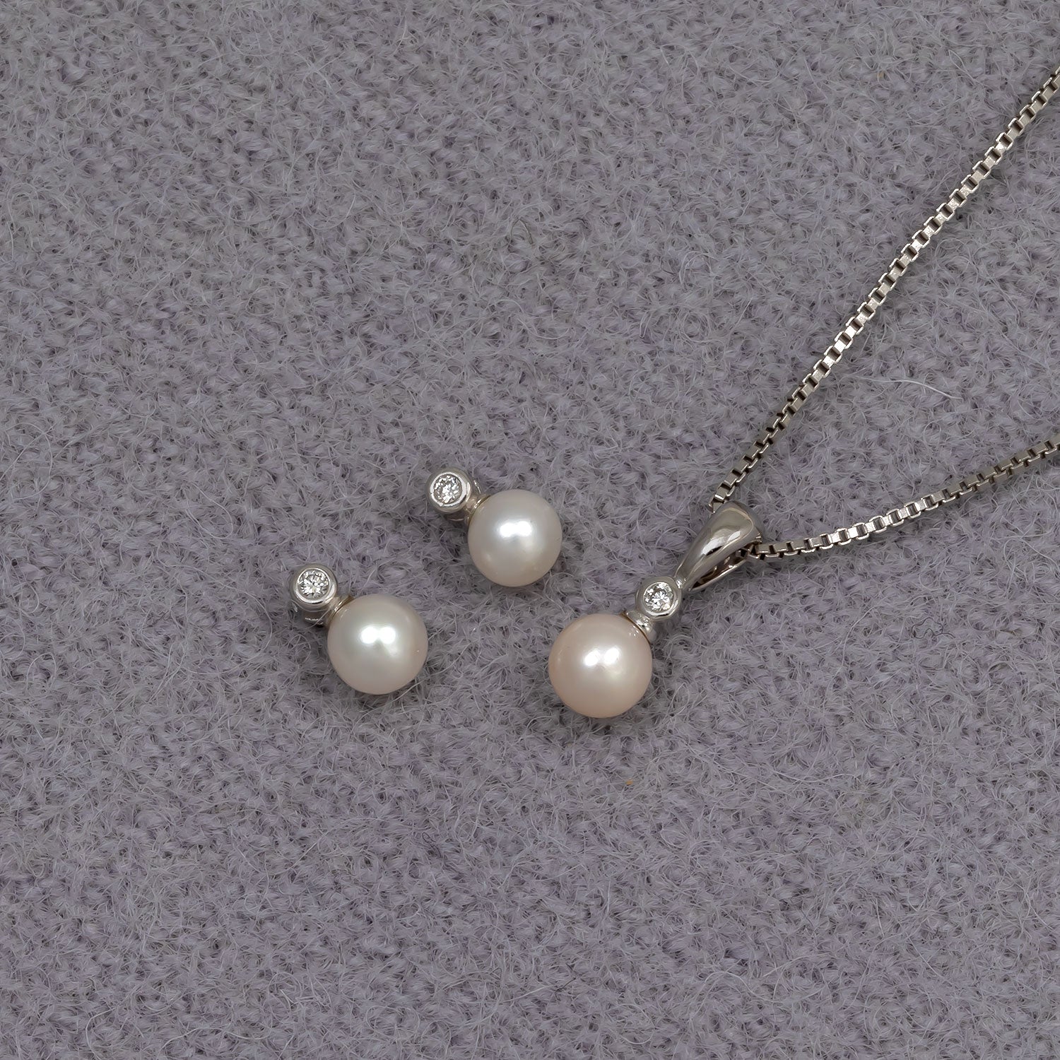 Pair of sterling silver stud earrings with freshwater pearls and small lab-grown diamonds, alongside a matching pendant necklace, displayed on soft grey fabric.