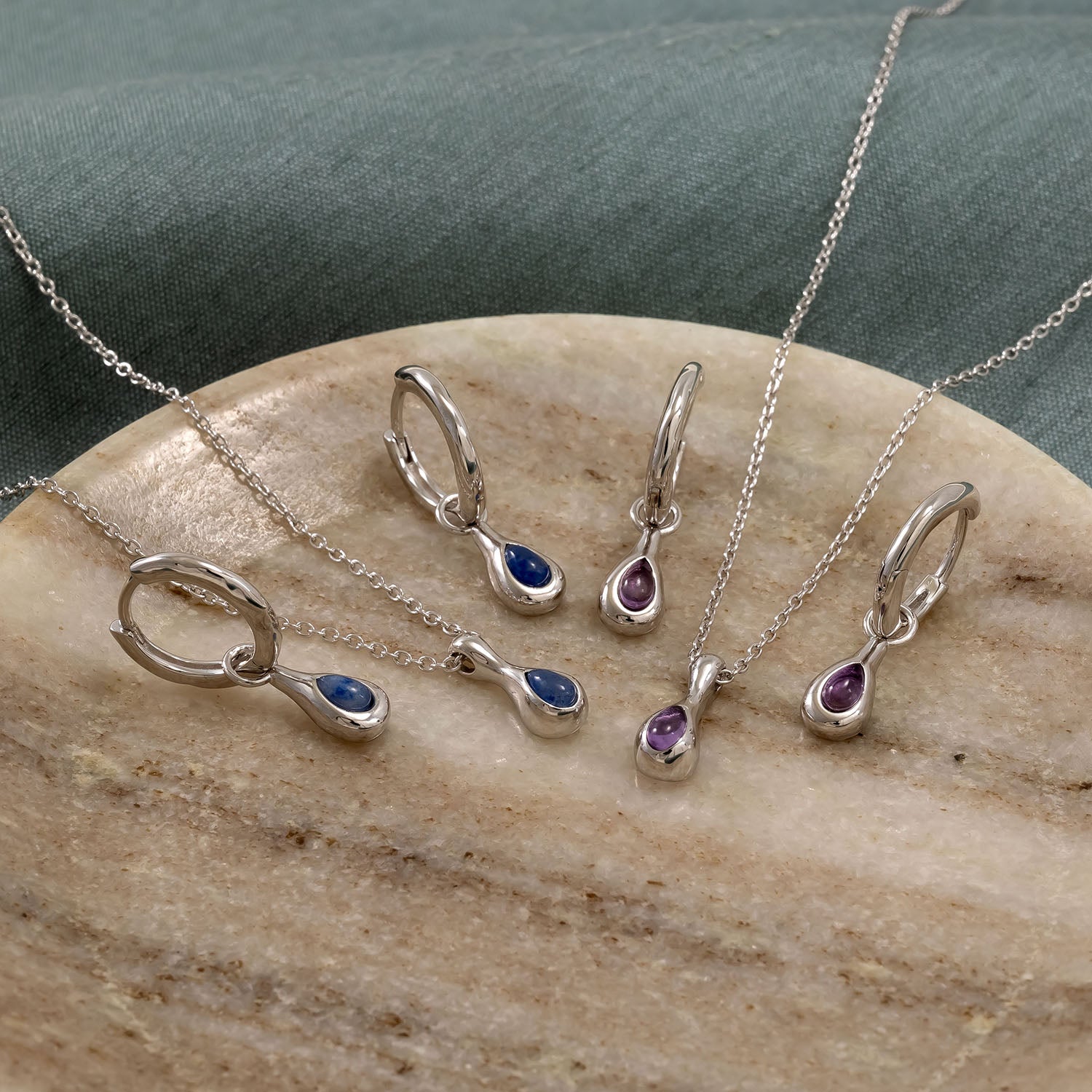 Sterling silver hinge hoop earrings and matching pendants with teardrop-shaped amethyst and blue gemstones, displayed on a smooth stone surface with a soft fabric background.