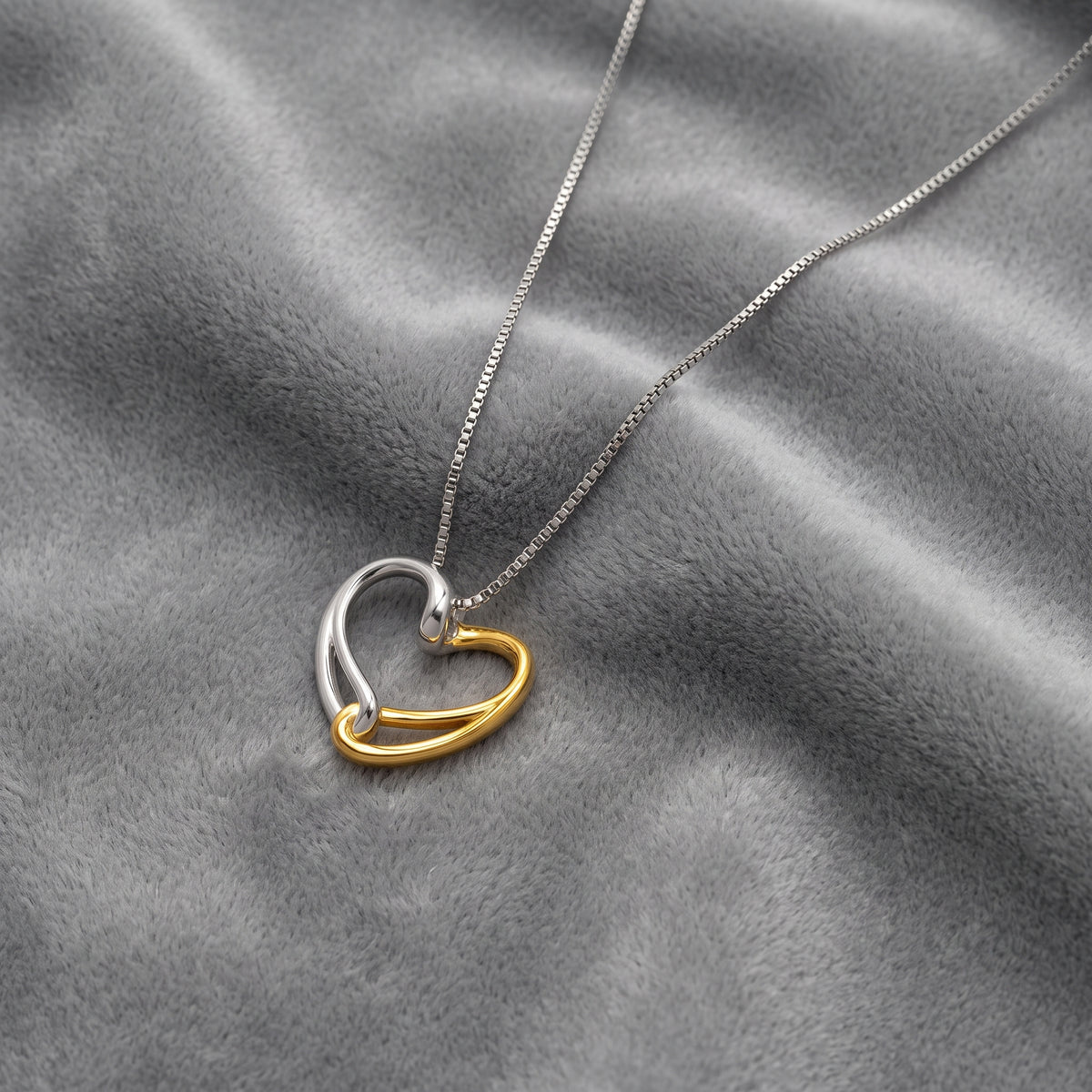 A silver chain with a two-tone heart-shaped pendant, featuring one half in polished silver and the other in polished yellow gold vermeil, resting on a textured gray fabric background.
