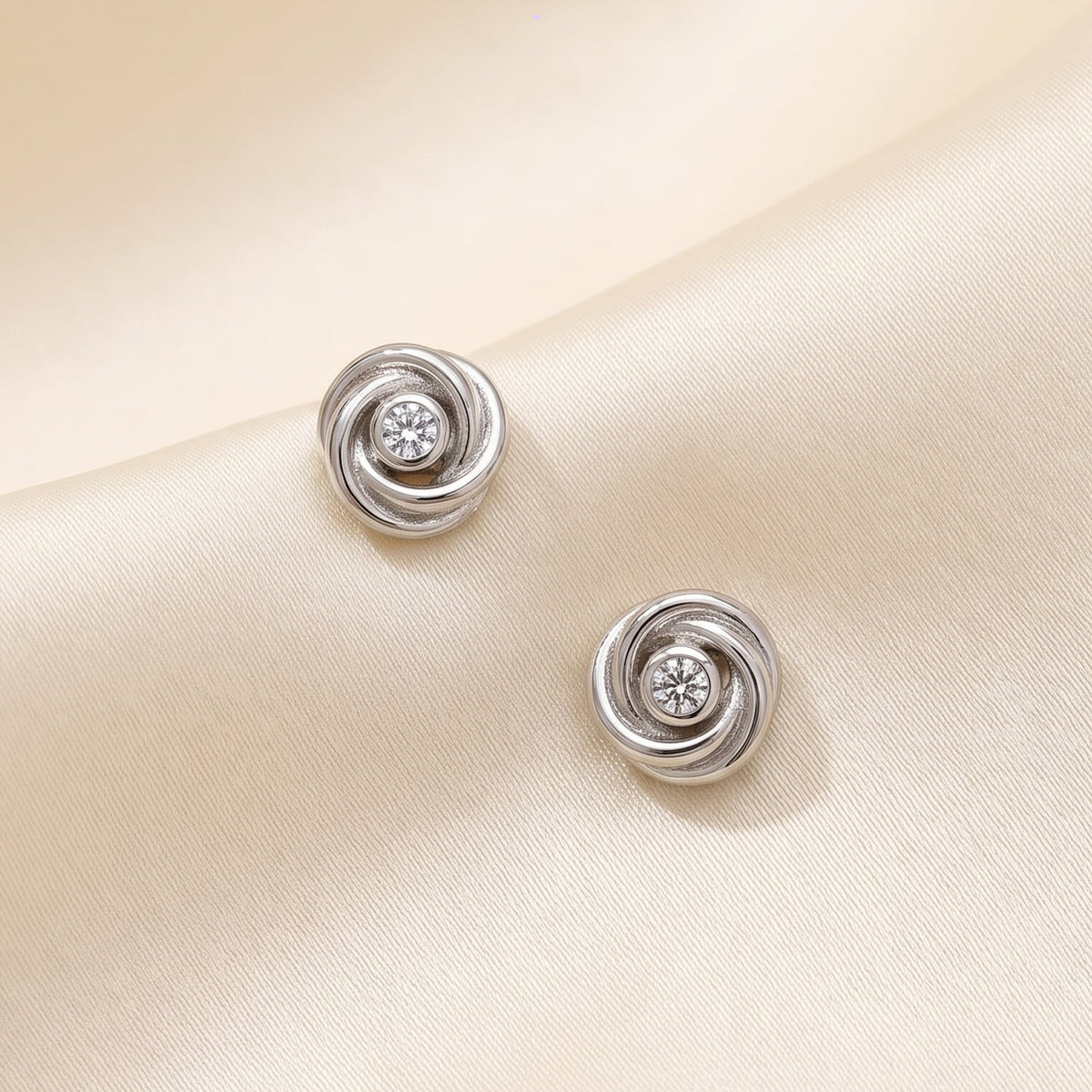 Entangled Silver & 0.13 Carat Lab Grown Diamond Love Knot Stud Earrings displayed on a cream silk fabric surface, showcasing their intricate knot design and sparkling center stones.