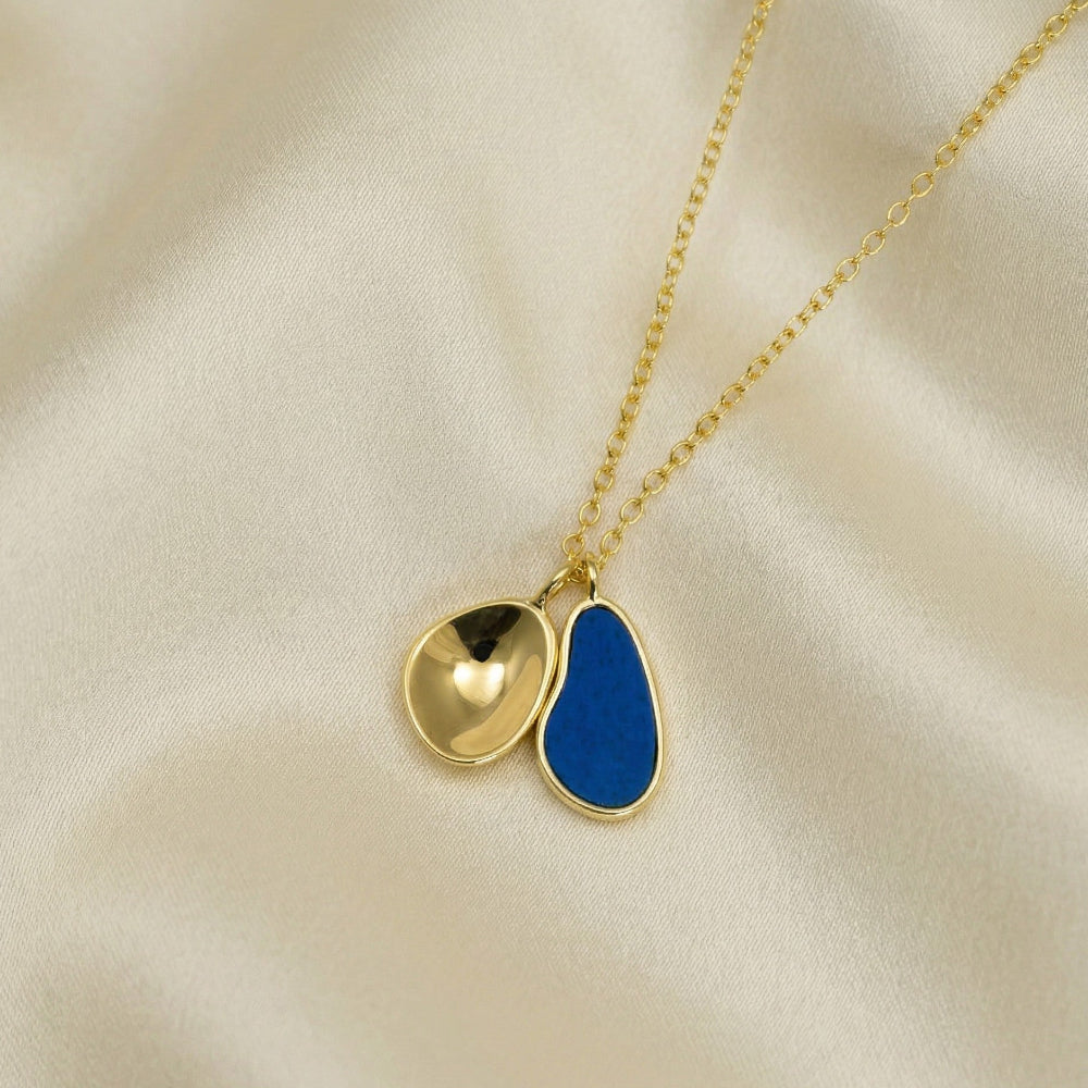 Elegant gold vermeil necklace featuring a double pendant with a polished sculptural charm and a deep blue lapis stone, displayed on a soft cream fabric background.