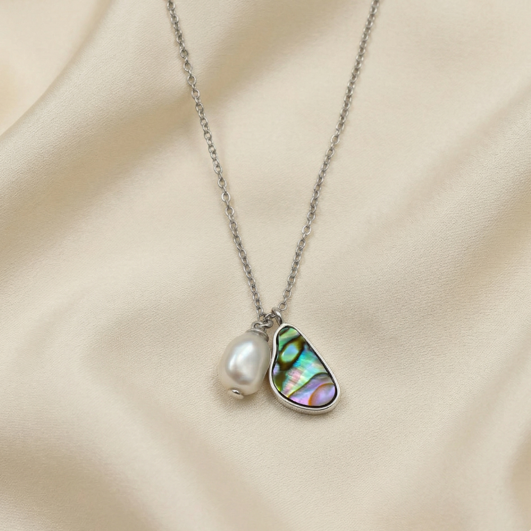 Delicate sterling silver necklace featuring a double pendant with a luminous baroque pearl and an iridescent abalone shell set in a sculptural silver frame, displayed on soft beige fabric.