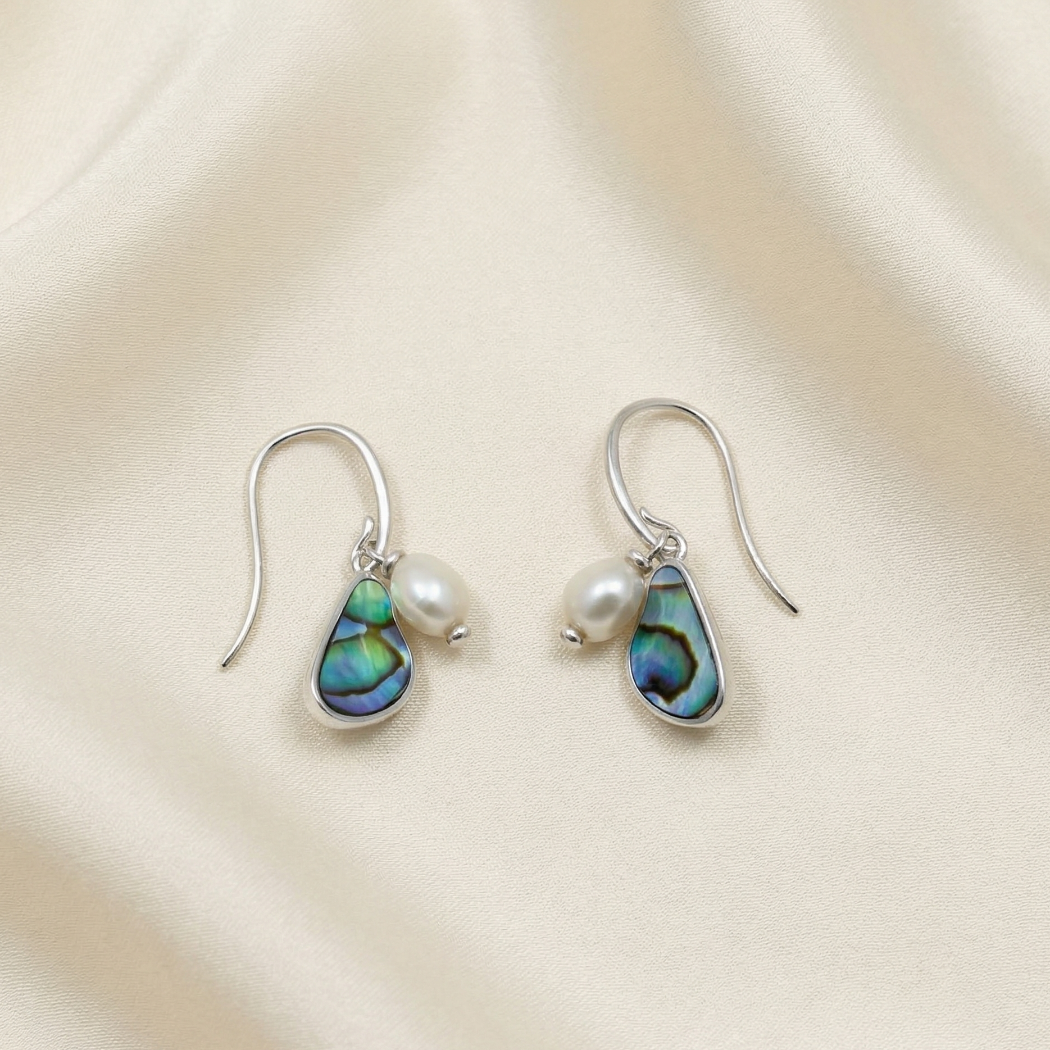 Pair of sterling silver drop earrings featuring irregular baroque freshwater pearls and teardrop-shaped abalone shells, displayed on a soft cream fabric background.