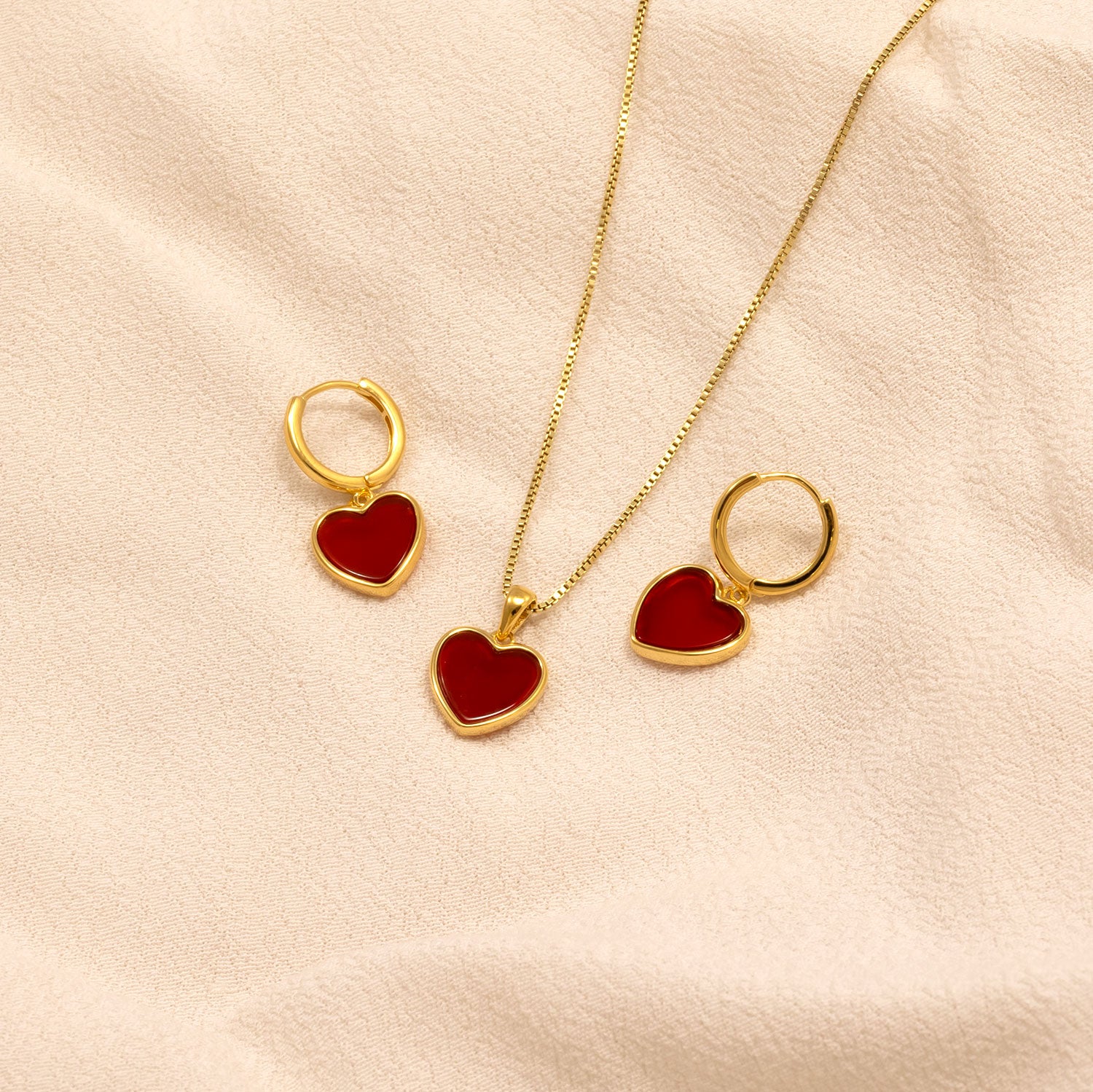 Set of gold vermeil hoop earrings and a matching pendant necklace featuring vibrant red agate heart-shaped charms, displayed on a soft beige fabric background.