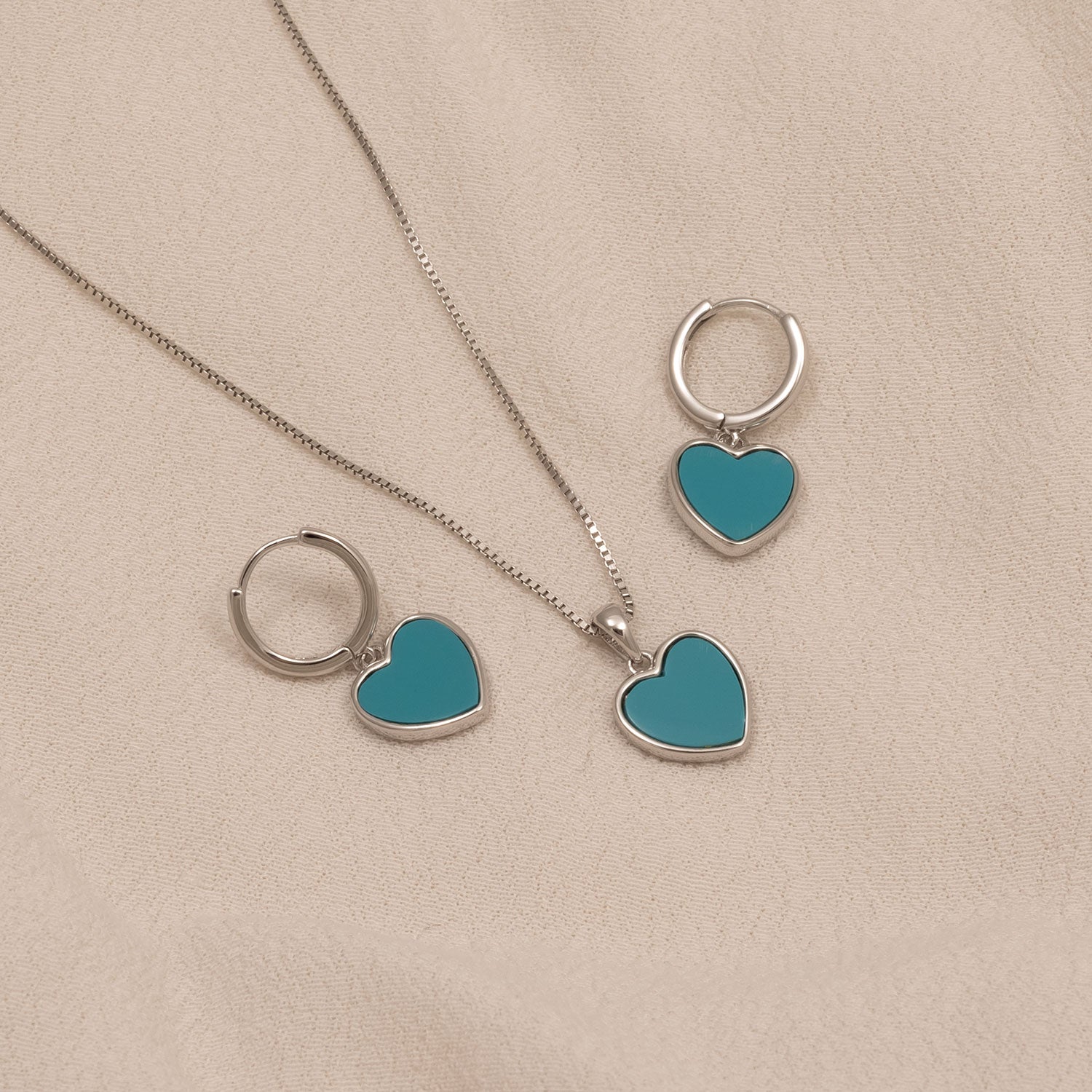 Pair of sterling silver hoop earrings and matching pendant necklace, each featuring turquoise heart-shaped charms resting on a soft beige fabric background.