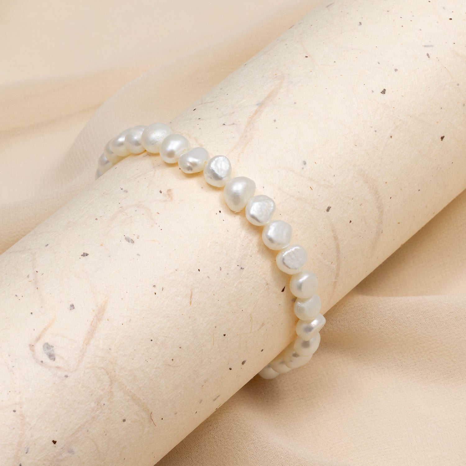 Delicate stretch bracelet featuring lustrous white freshwater pearls with organic shapes, displayed on a light, textured neutral fabric background.