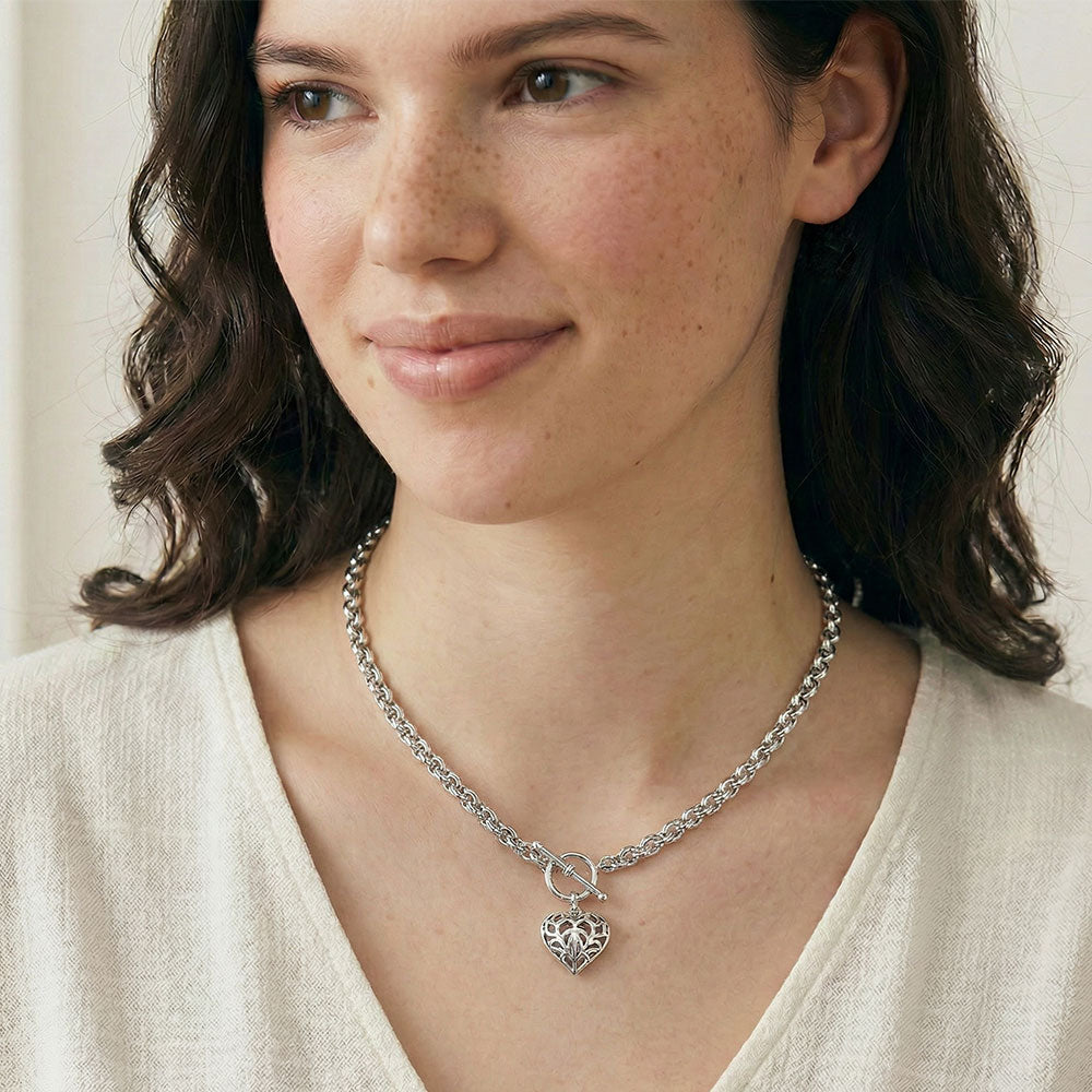 Close-up of a woman with dark wavy hair wearing a textured silver chain necklace featuring an intricate heart-shaped pendant, dressed in a soft cream top.