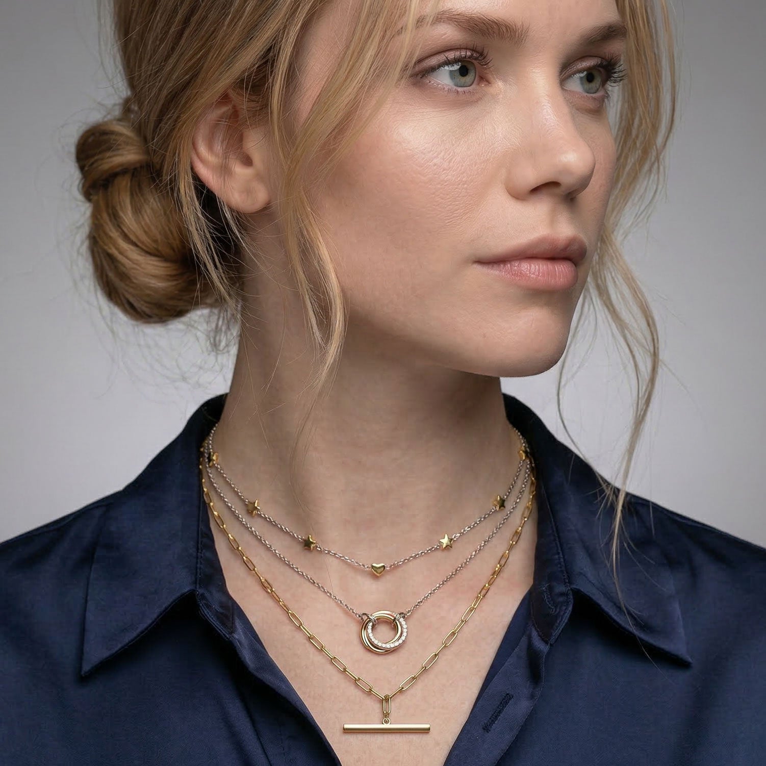 Profile view of a woman wearing layered gold vermeil necklaces with delicate chain designs, including a small heart charm, a circular pendant, and a bar pendant, styled with a dark blouse.