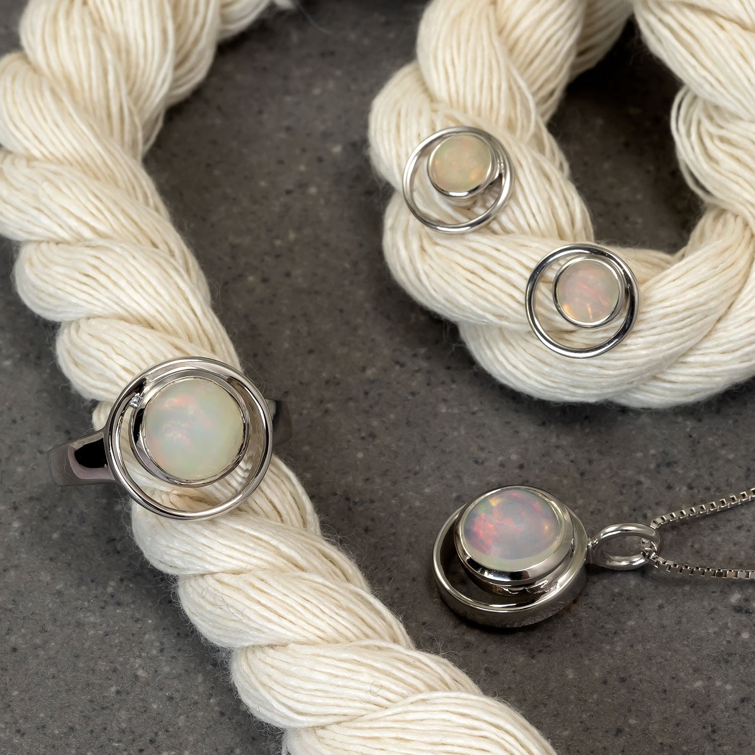 Sterling silver ring and matching jewellery set featuring luminous opal stones encased in polished circular silver frames, displayed on soft cream yarn against a dark surface.