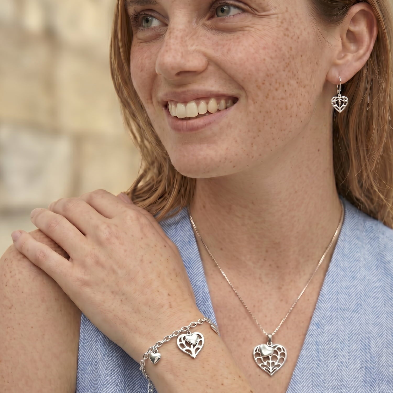 A smiling woman wearing a silver heart pendant necklace, matching heart drop earrings, and a silver heart bracelet, shown in a close-up lifestyle shot.