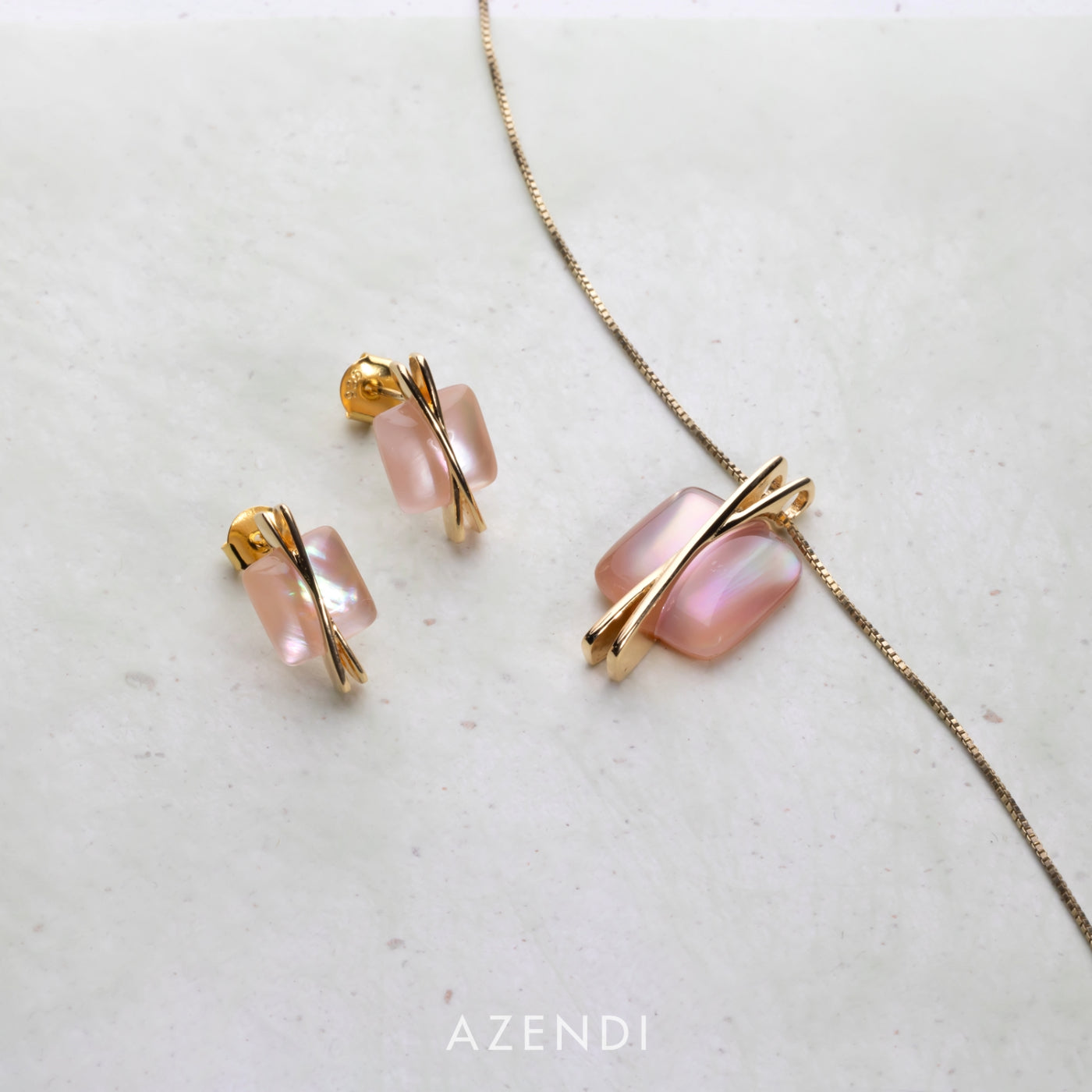 A pair of gold vermeil earrings and matching pendant featuring square-shaped pink mother of pearl stones accented by sleek crossover bars, displayed on a soft white background.