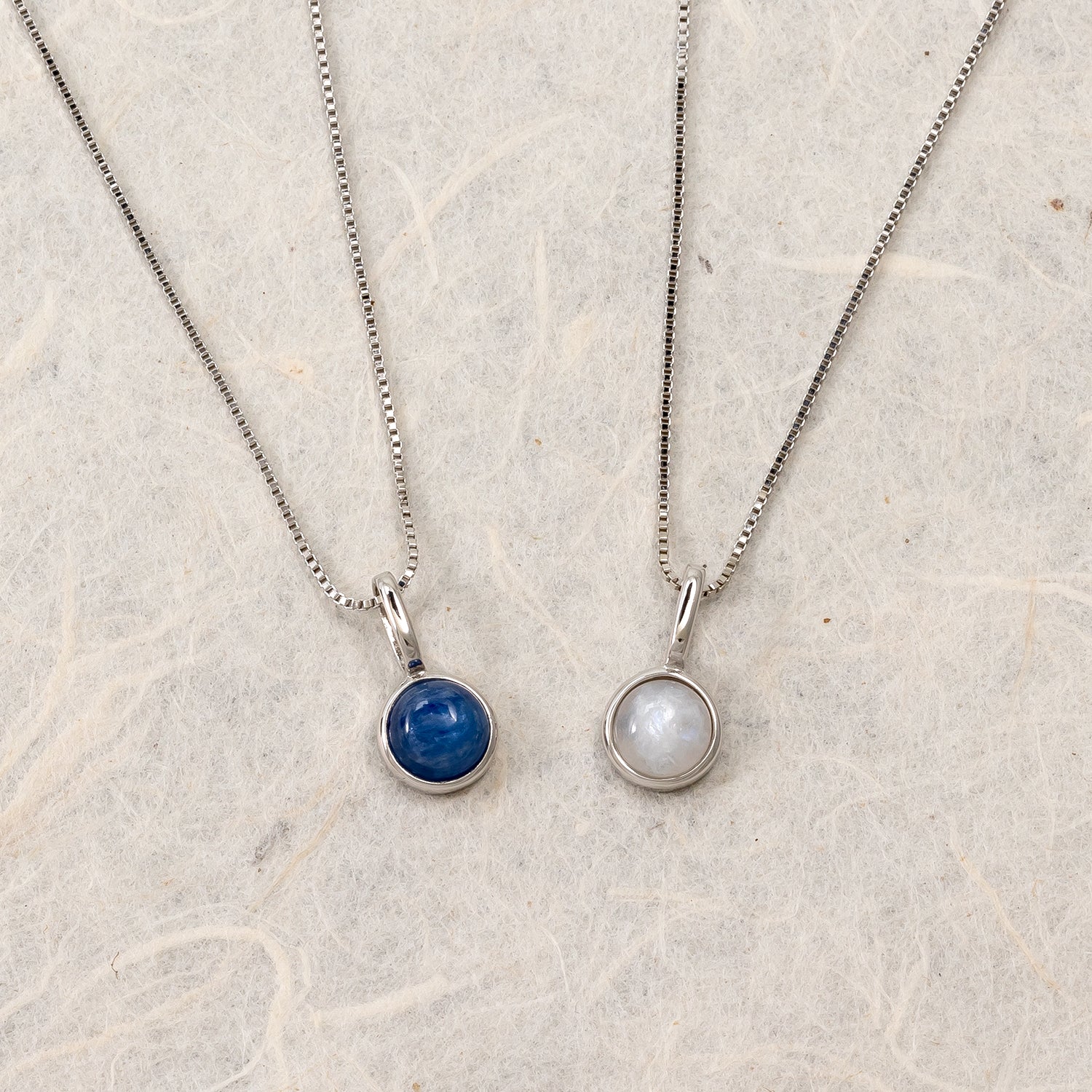 Two sterling silver necklaces each featuring a round bezel-set gemstone pendant, one with a deep blue stone and the other showcasing a luminous moonstone, on fine chains.