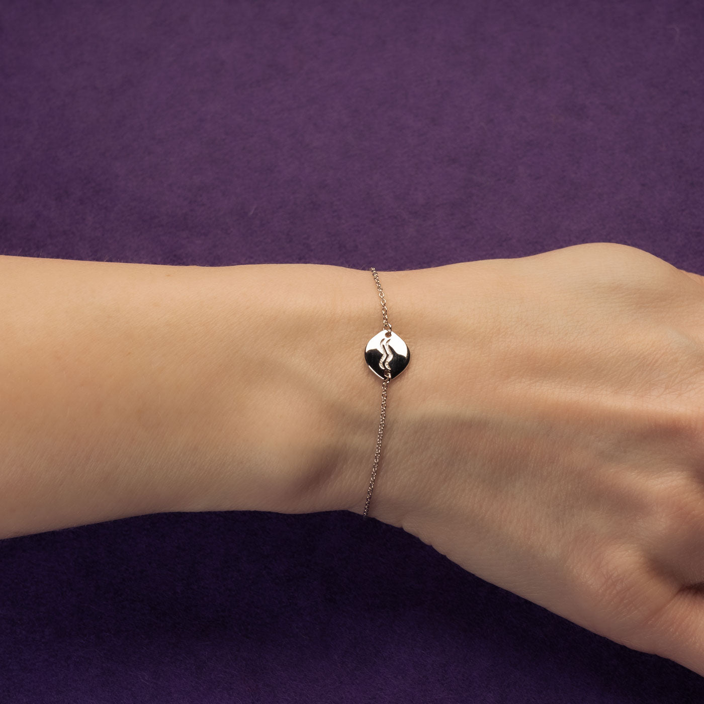 Delicate sterling silver bracelet featuring a polished Aquarius symbol charm on a fine adjustable chain, styled on the wrist against a rich purple background.