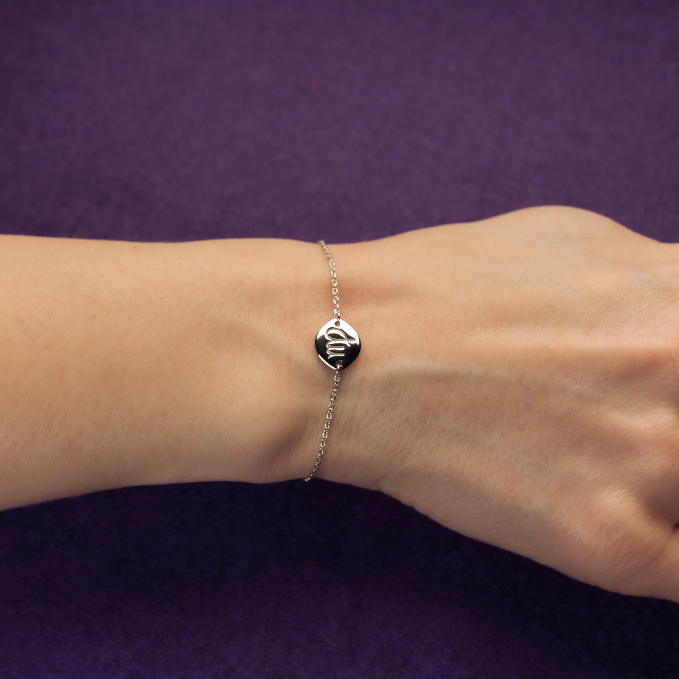 Delicate sterling silver adjustable bracelet featuring a polished round charm engraved with the Virgo zodiac symbol, showcased on a wrist against a deep purple background.