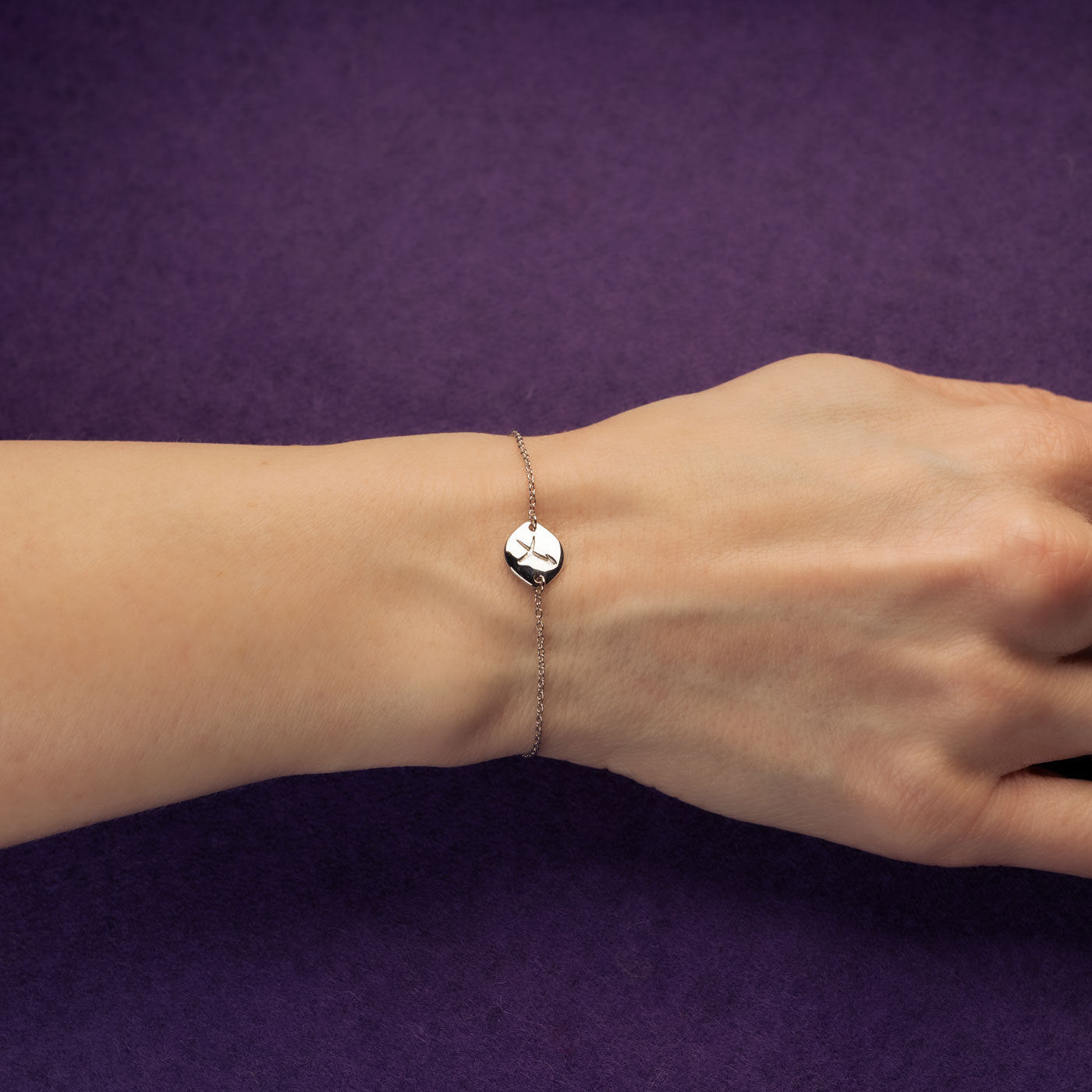 Delicate sterling silver adjustable chain bracelet featuring a subtle Sagittarius zodiac symbol charm, styled on a wrist against a rich purple background.
