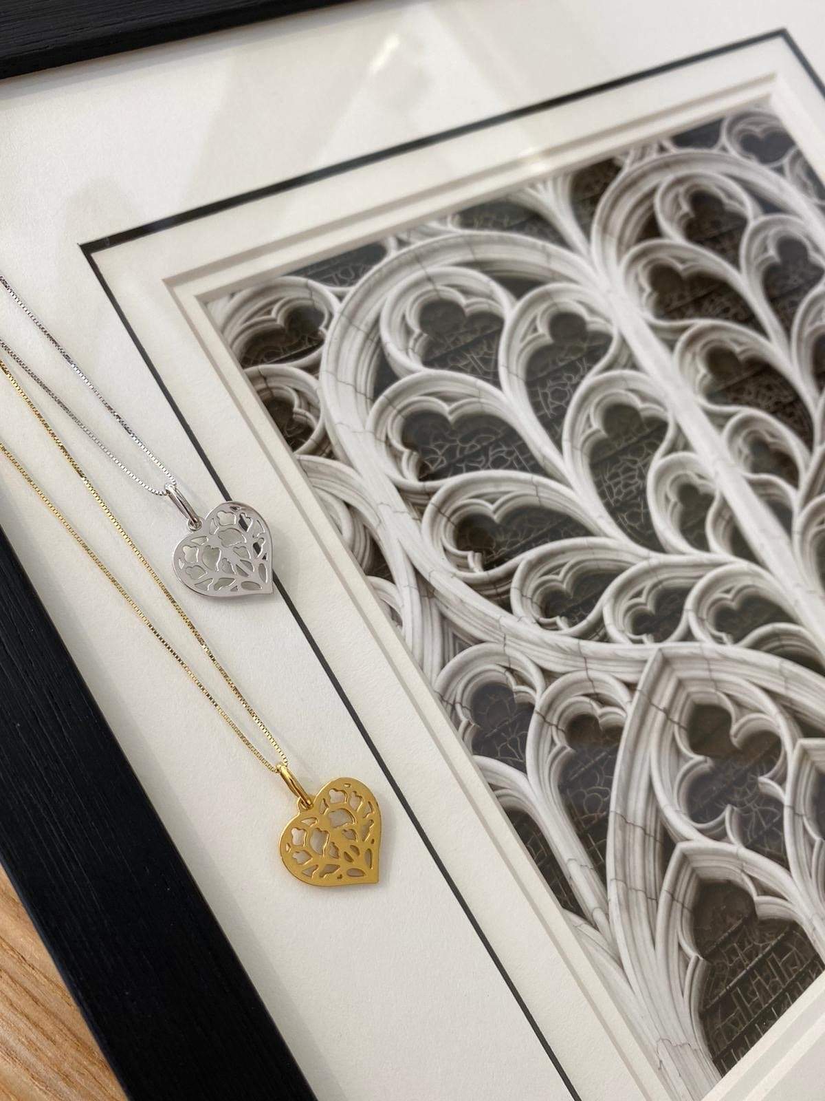 Two heart-shaped pendants with an open filigree design, one in 9 carat white gold and the other in yellow gold, displayed on delicate chains over a framed architectural detail.