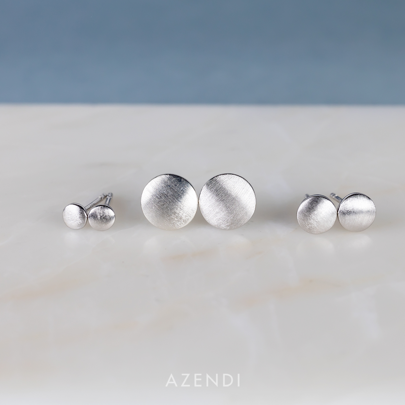 Set of three pairs of sterling silver stud earrings with a double curved, satin brushed finish, displayed on a smooth marble surface against a muted blue backdrop.