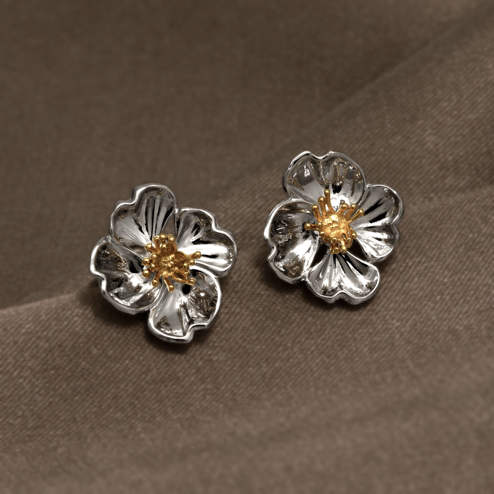 Pair of sterling silver earrings shaped as sculptural primrose flowers with detailed gold vermeil centers, presented on a soft brown fabric background.