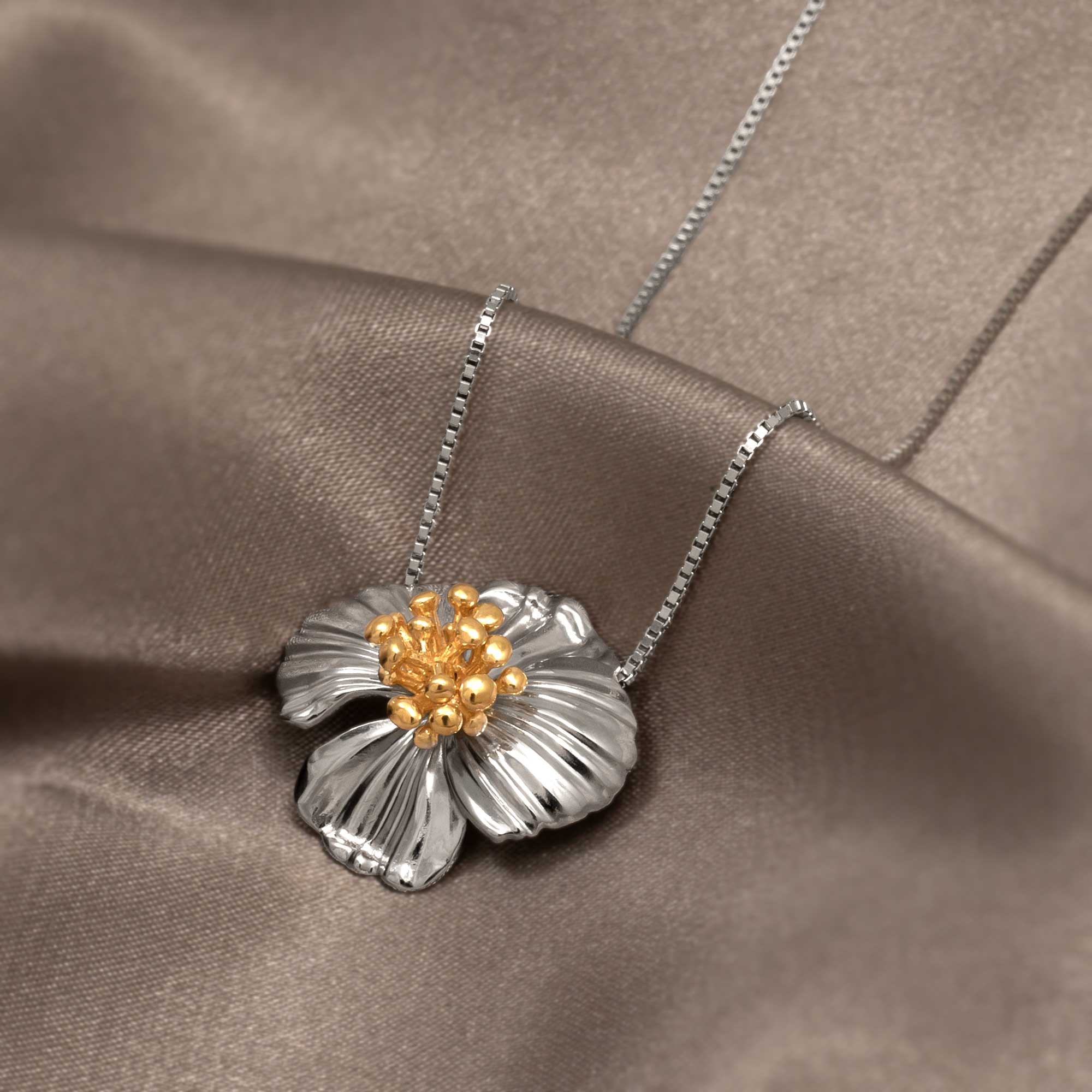 Sterling silver pendant featuring a sculpted poppy flower with gold vermeil stamen details, displayed on a fine chain over smooth taupe fabric for a refined finish.