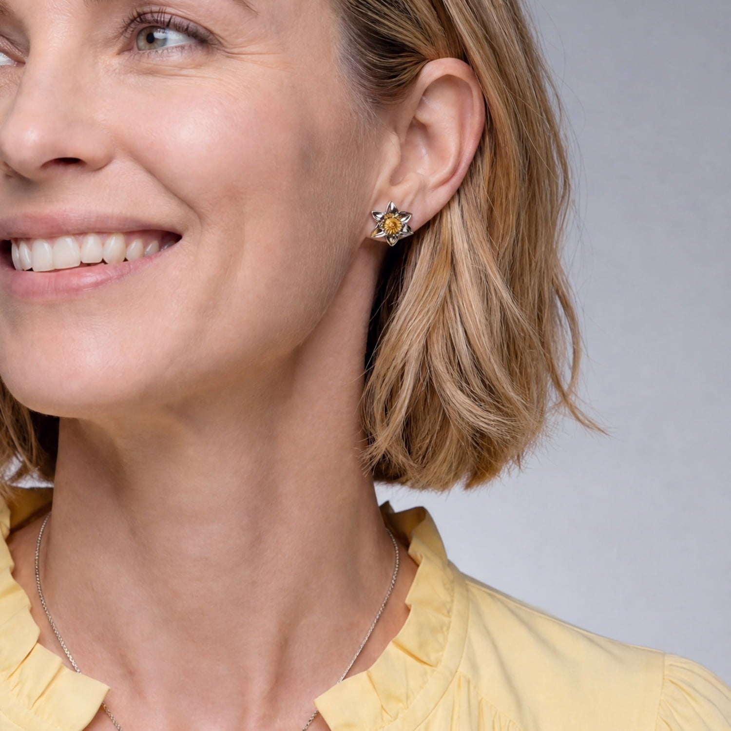 Sterling silver stud earrings inspired by daffodil blooms, featuring detailed petals and a central yellow gemstone, elegantly worn on the ear.