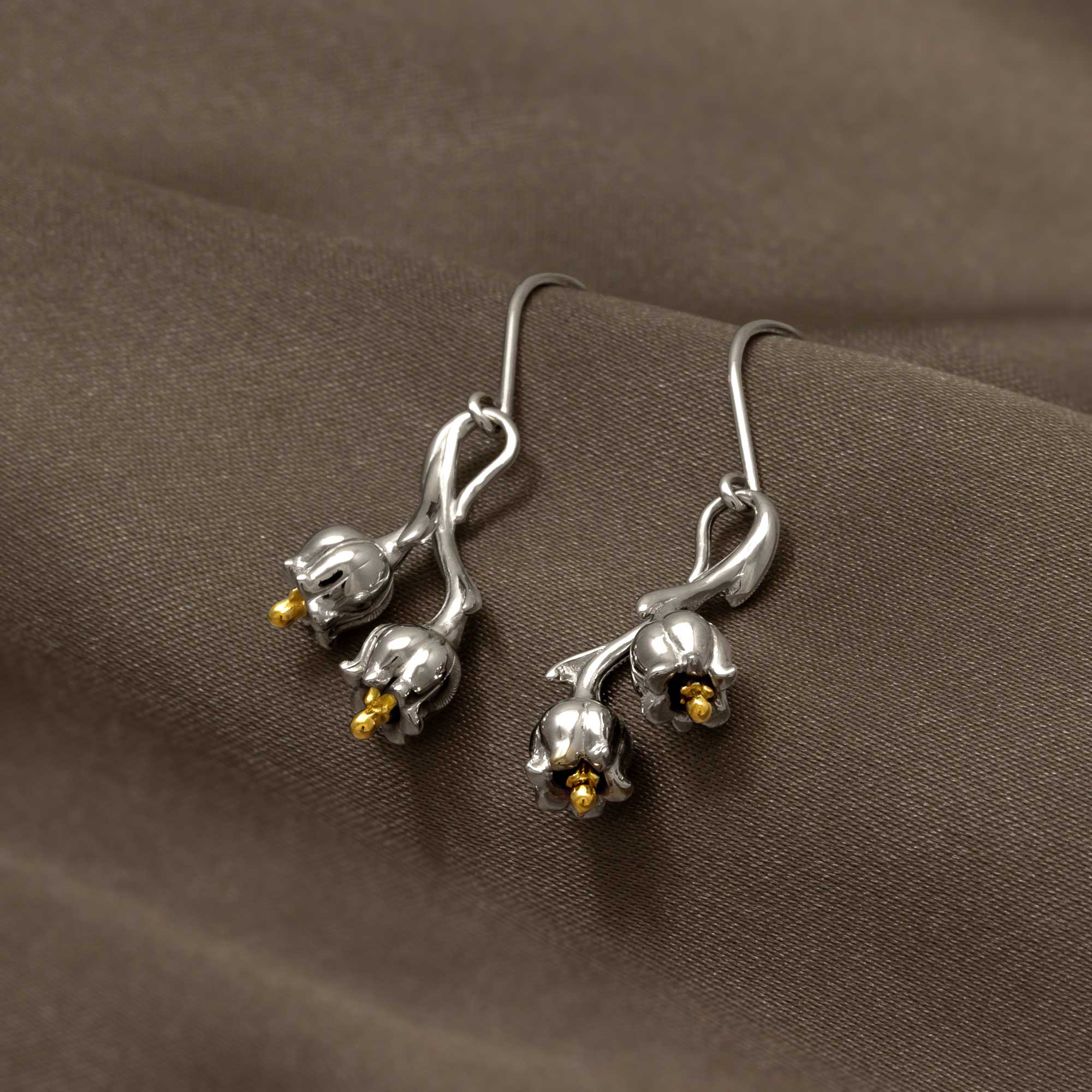 Sterling silver earrings featuring sculptural lily of the valley flowers with delicate gold vermeil accents, elegantly displayed on soft brown fabric.