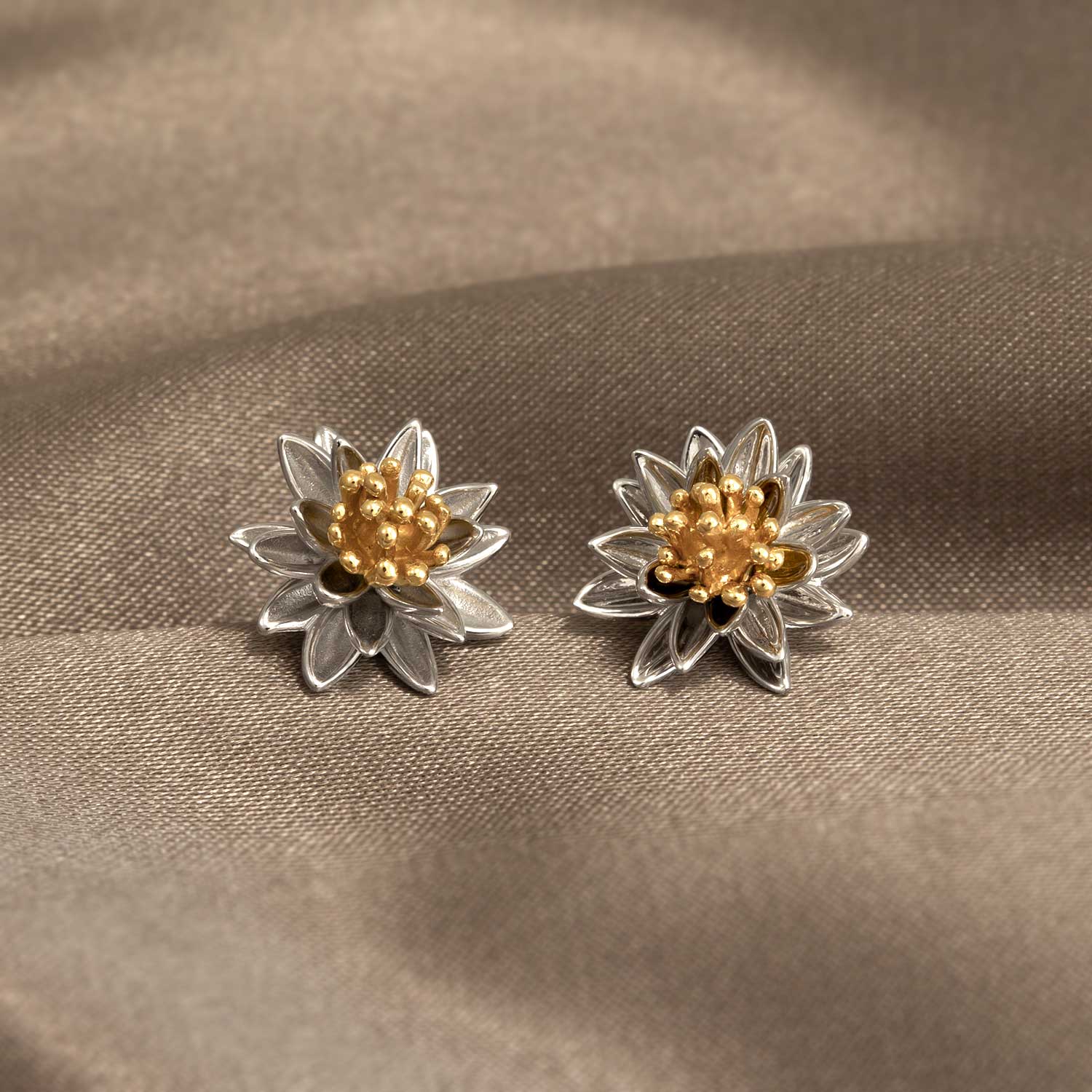 Sterling silver earrings designed as sculptural water lily flowers with textured gold vermeil centers, showcasing expert British craftsmanship on a soft neutral fabric background.