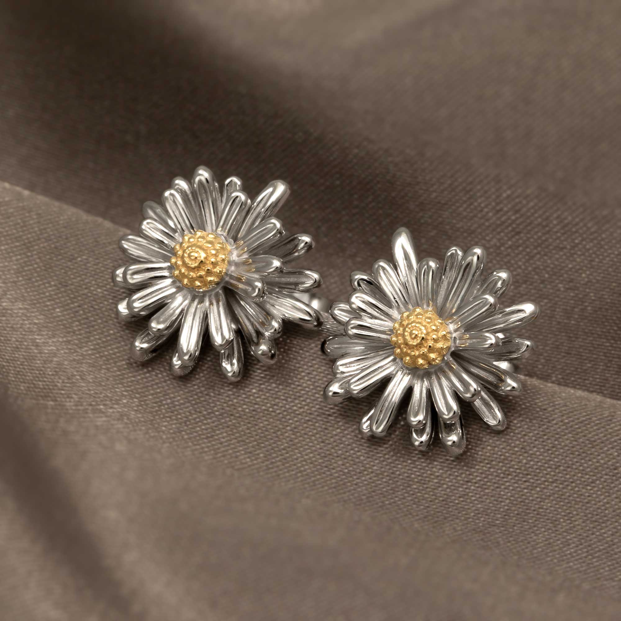Pair of sterling silver earrings designed as sculptural daisies with textured gold vermeil centers, displayed on smooth taupe fabric highlighting fine British craftsmanship.
