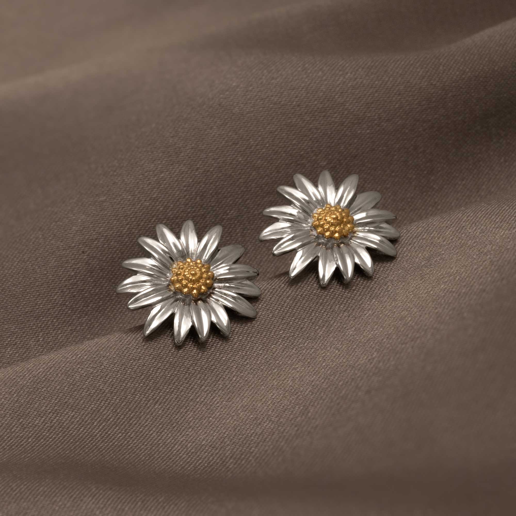 Pair of sterling silver earrings crafted as sculptural daisies with detailed petals and textured gold vermeil centers, displayed on soft brown fabric.