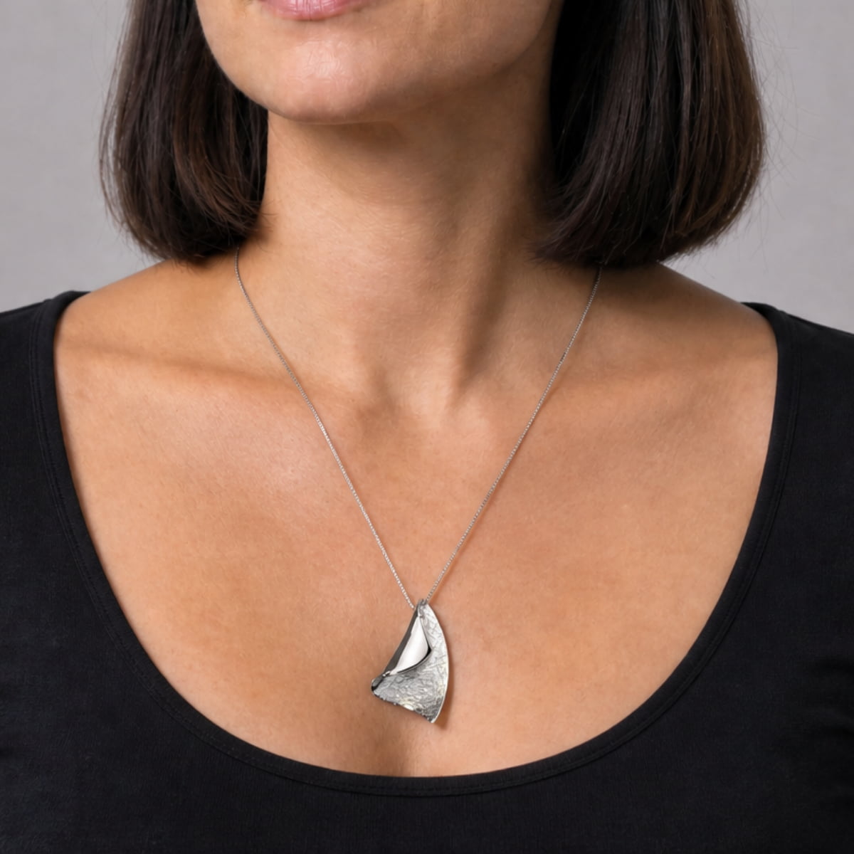 A close-up of a woman wearing a silver pendant necklace with an organic, wavy leaf-like shape over a black top.