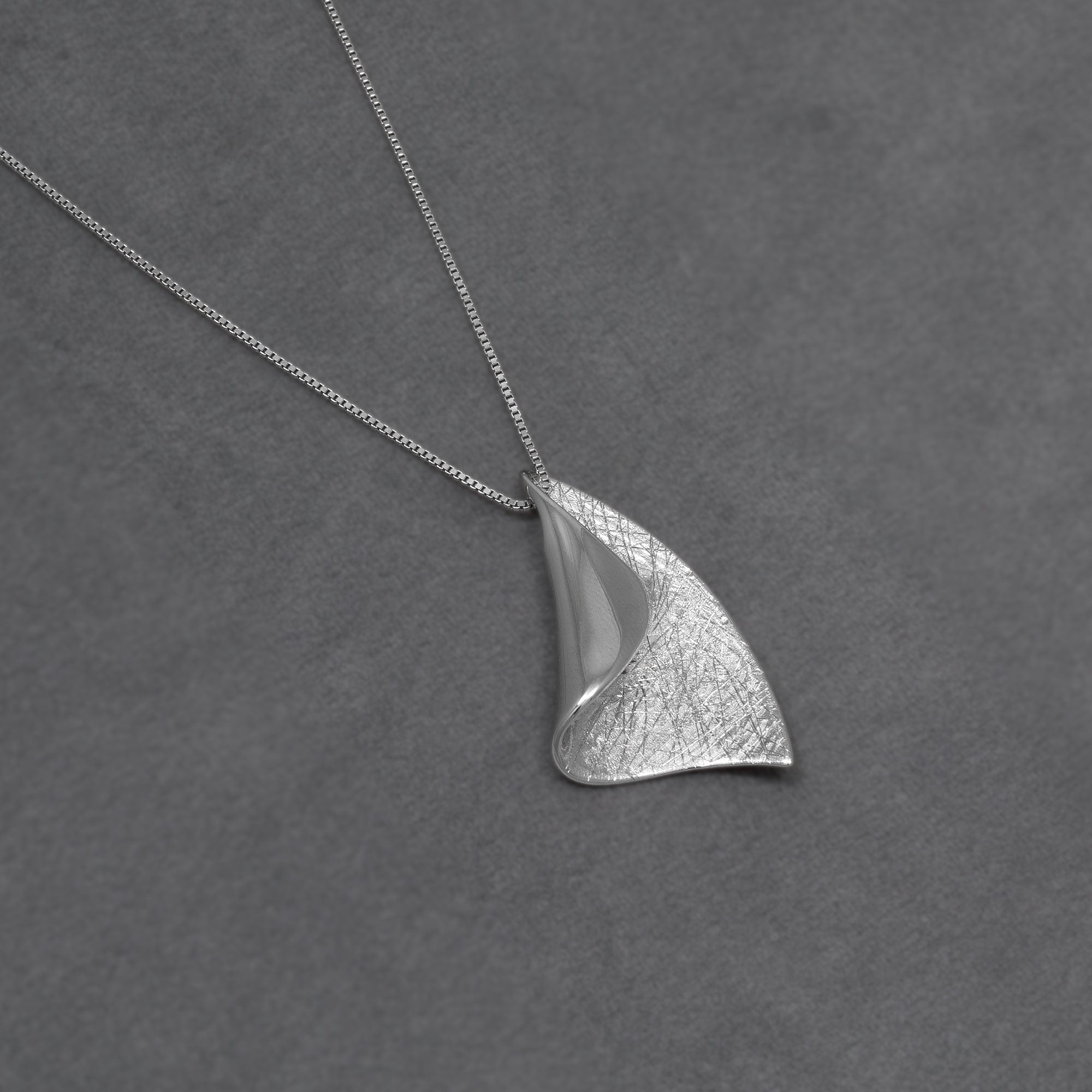 A silver pendant necklace featuring a textured, organic leaf-like shape on a delicate chain, displayed on a dark grey textured background.