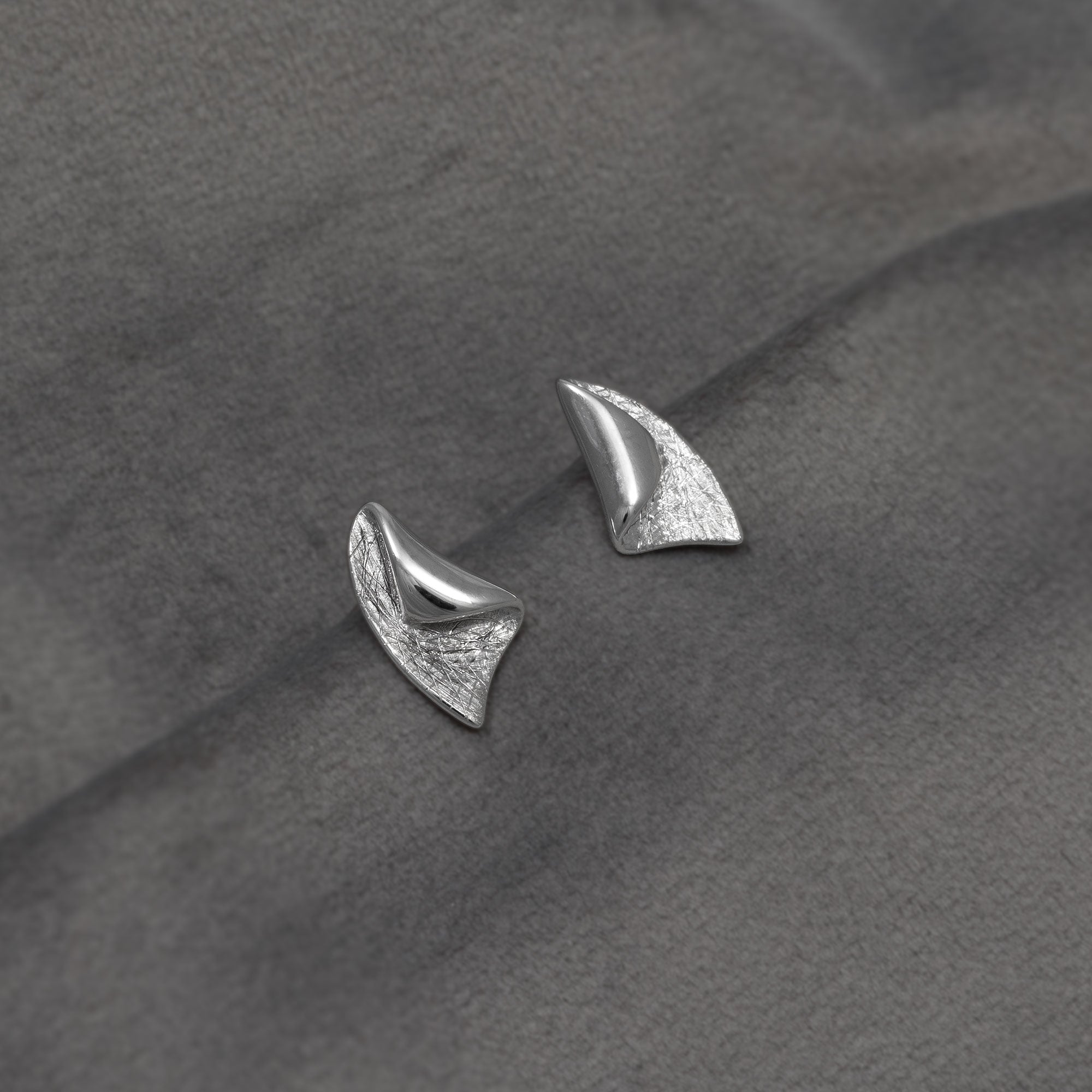 A pair of textured silver stud earrings with a curved, leaf-like design, displayed on a dark grey fabric background.
