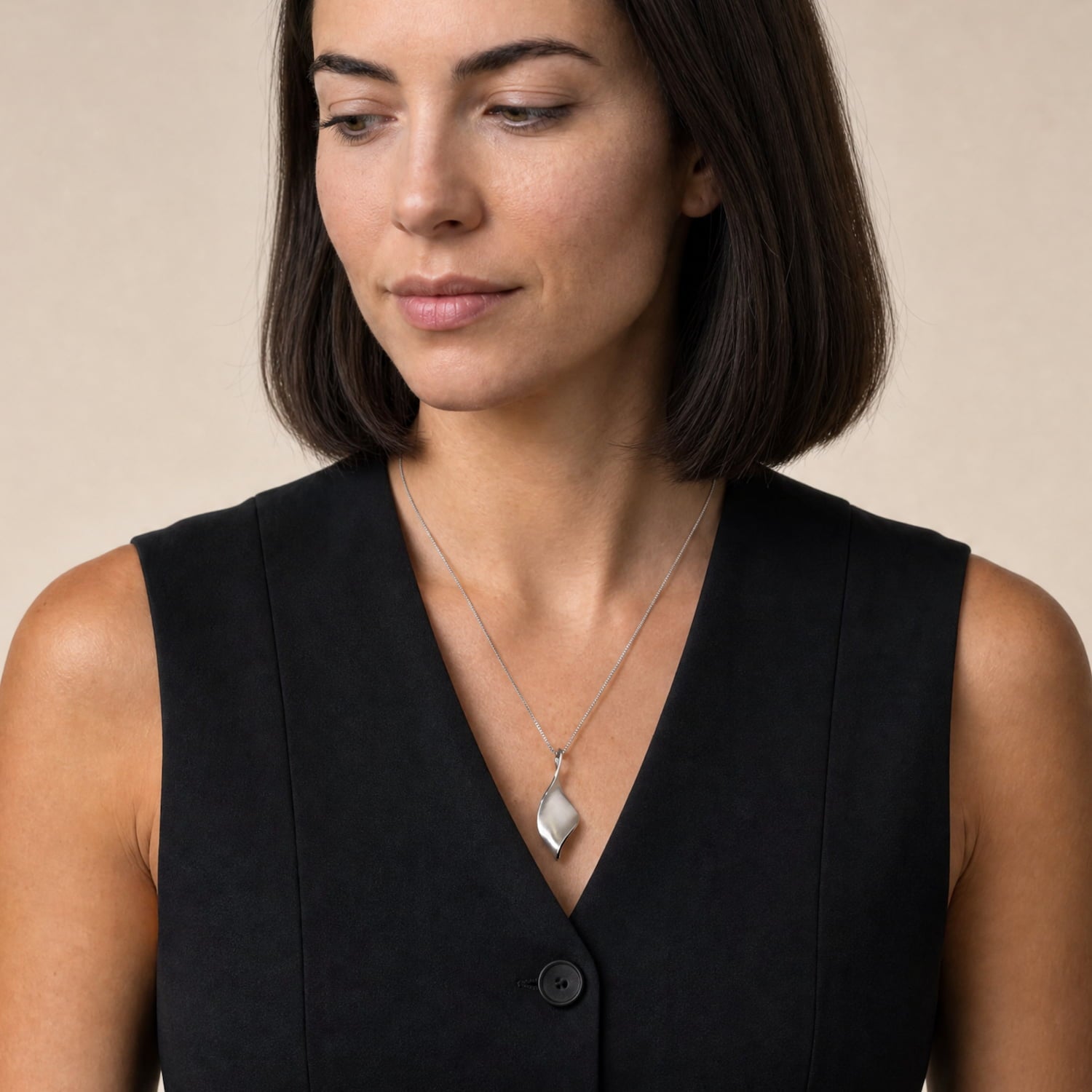 A woman with a short dark bob hairstyle wearing a twisting silver pendant, paired with a black sleeveless top, against a solid beige background.