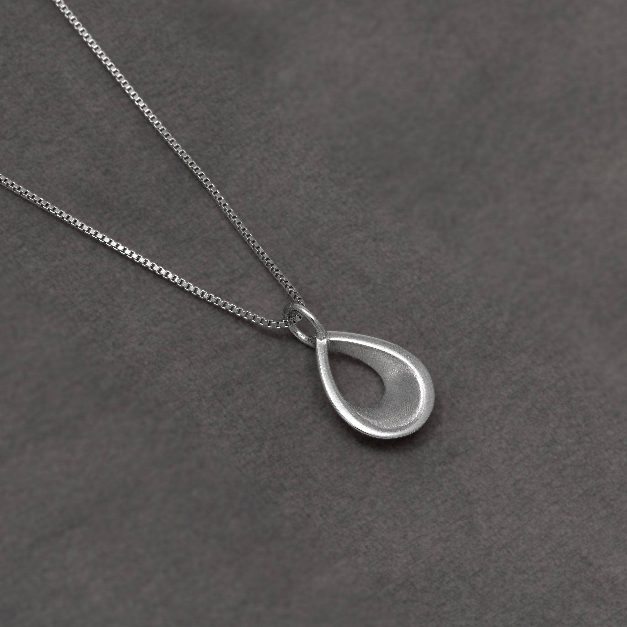 A silver teardrop pendant necklace on a delicate silver chain, displayed on a textured dark grey background.