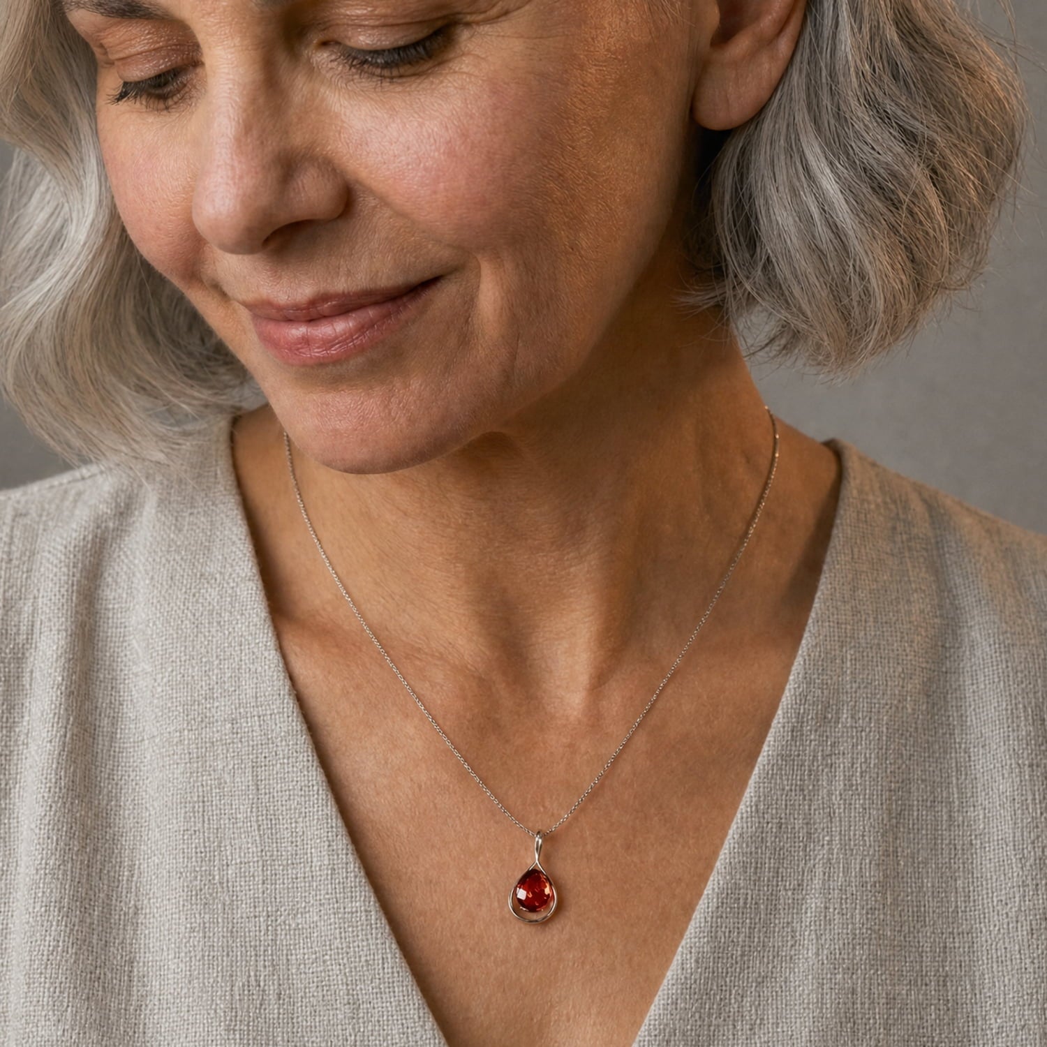 A close-up of a woman with short grey hair wearing a silver chain necklace with a teardrop-shaped amber gemstone pendant, paired with a light beige linen top.