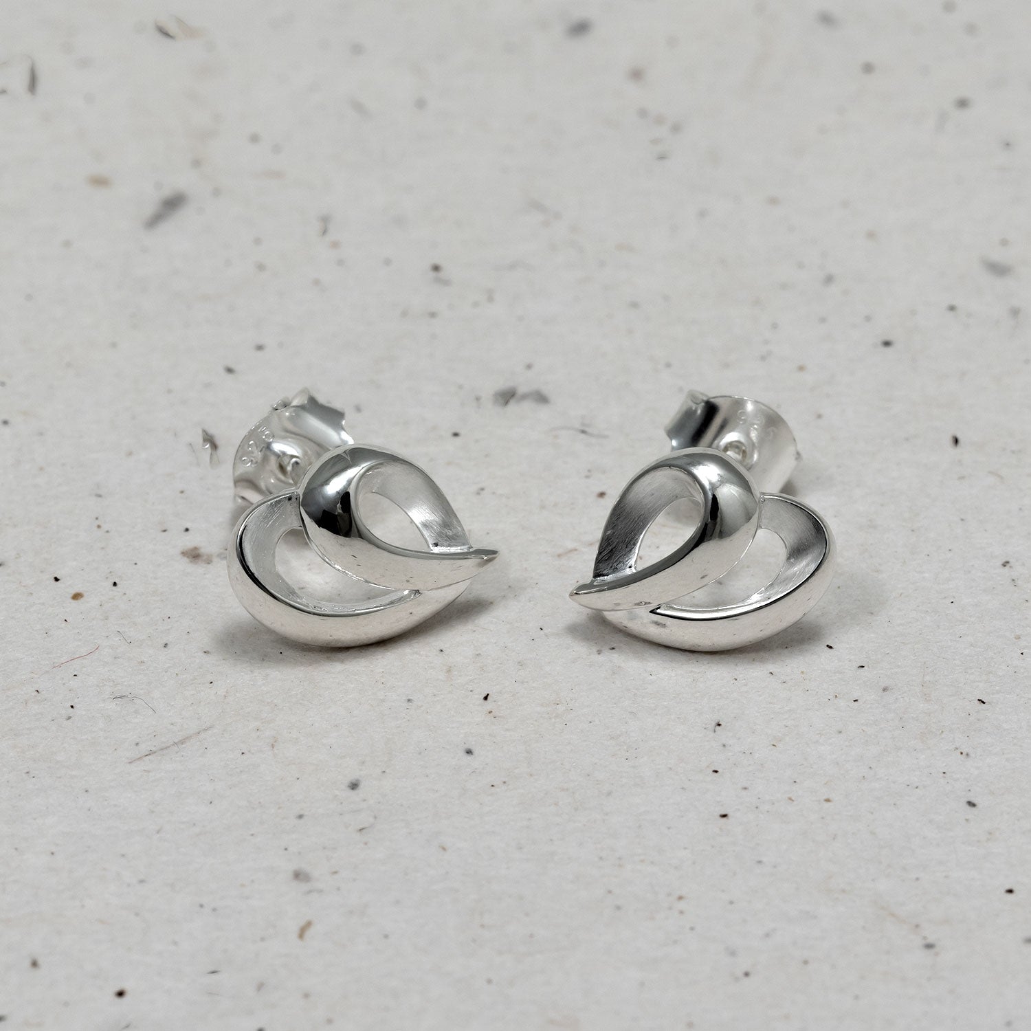 Sculptural sterling silver stud earrings shaped with flowing, intertwined curves forming a delicate heart-inspired design, reflecting refined contemporary silver jewellery.