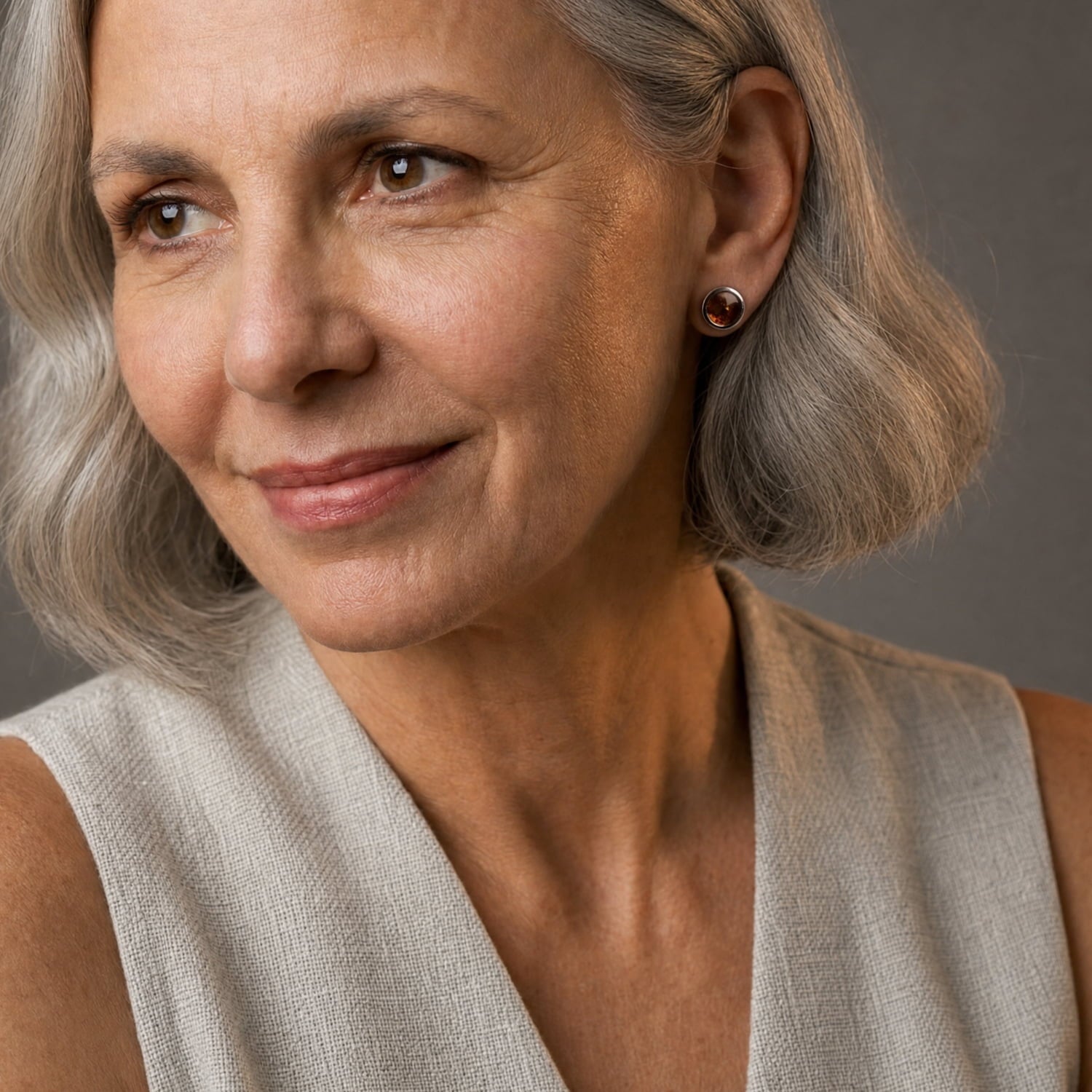 Close-up portrait of a woman with short grey hair wearing a small round stud earring with a dark amber gem, against a soft gray background.