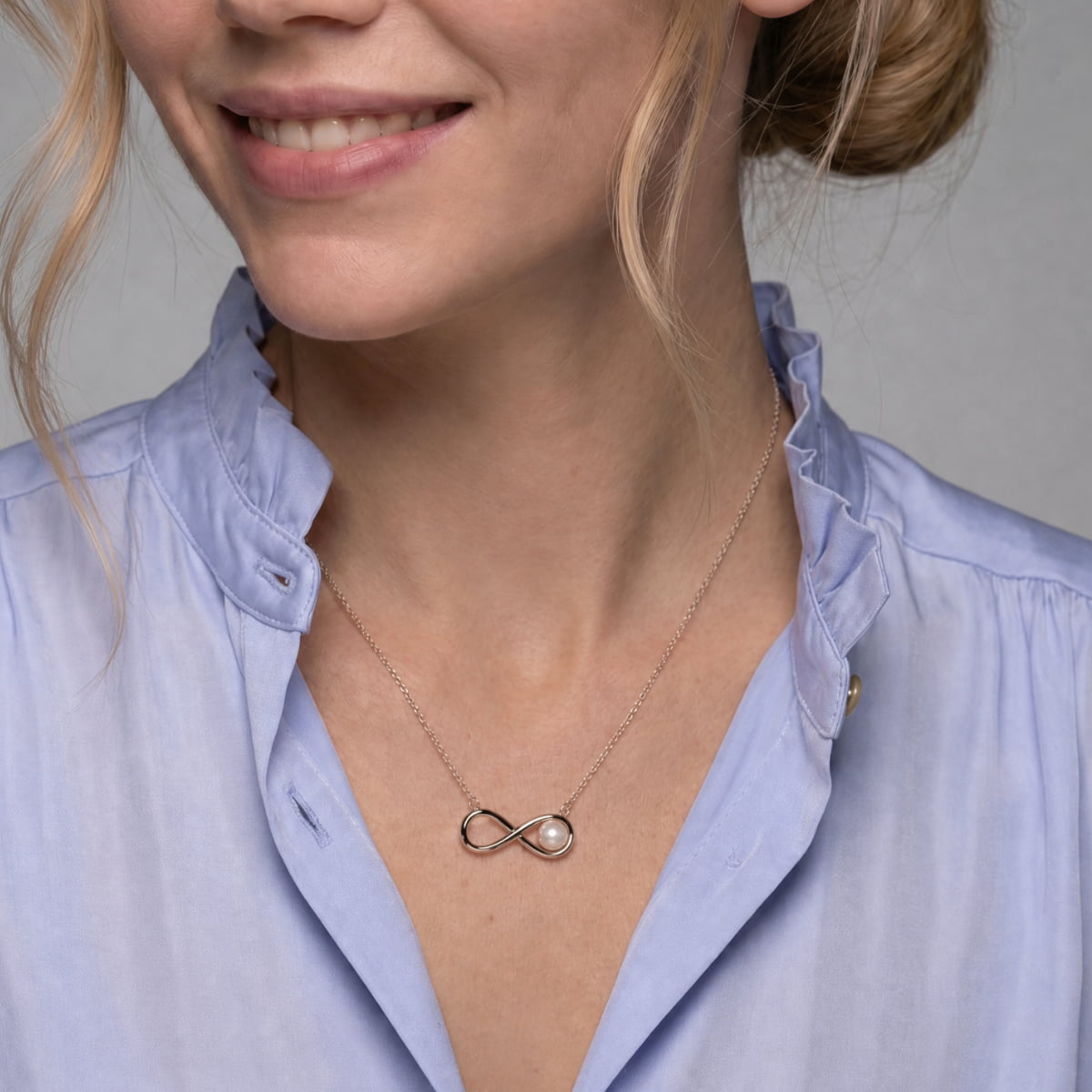 A close-up of a woman wearing a delicate silver infinity necklace with a freshwater pearl accent, styled with a light blue blouse.