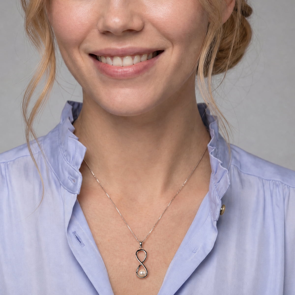 A close-up of a woman wearing a delicate silver infinity-shaped pendant necklace with a small white pearl at the bottom, styled with a light blue blouse.