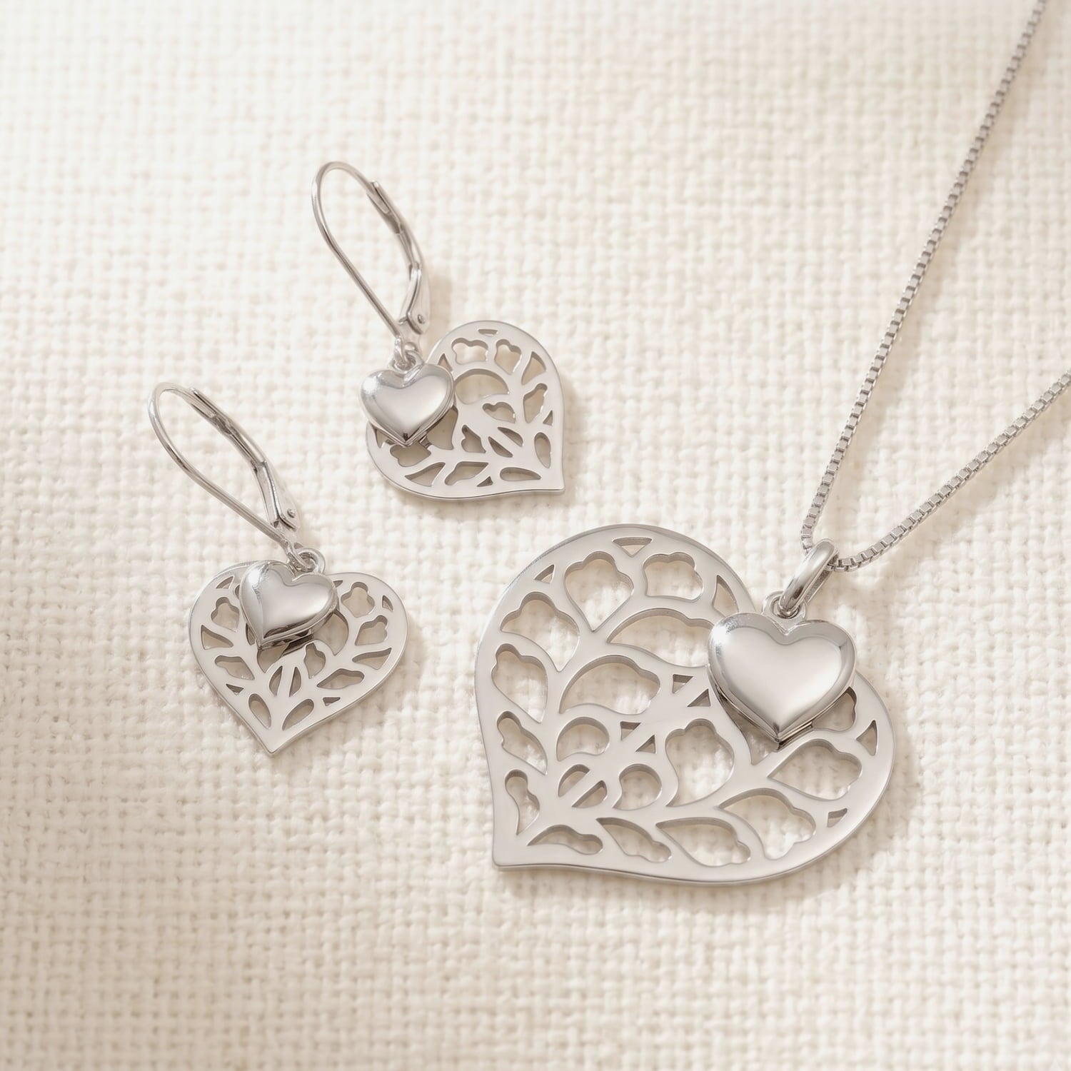 Sterling silver necklace and matching double drop earrings featuring intricate openwork hearts accented with smooth sculptural heart charms, displayed on a neutral fabric.