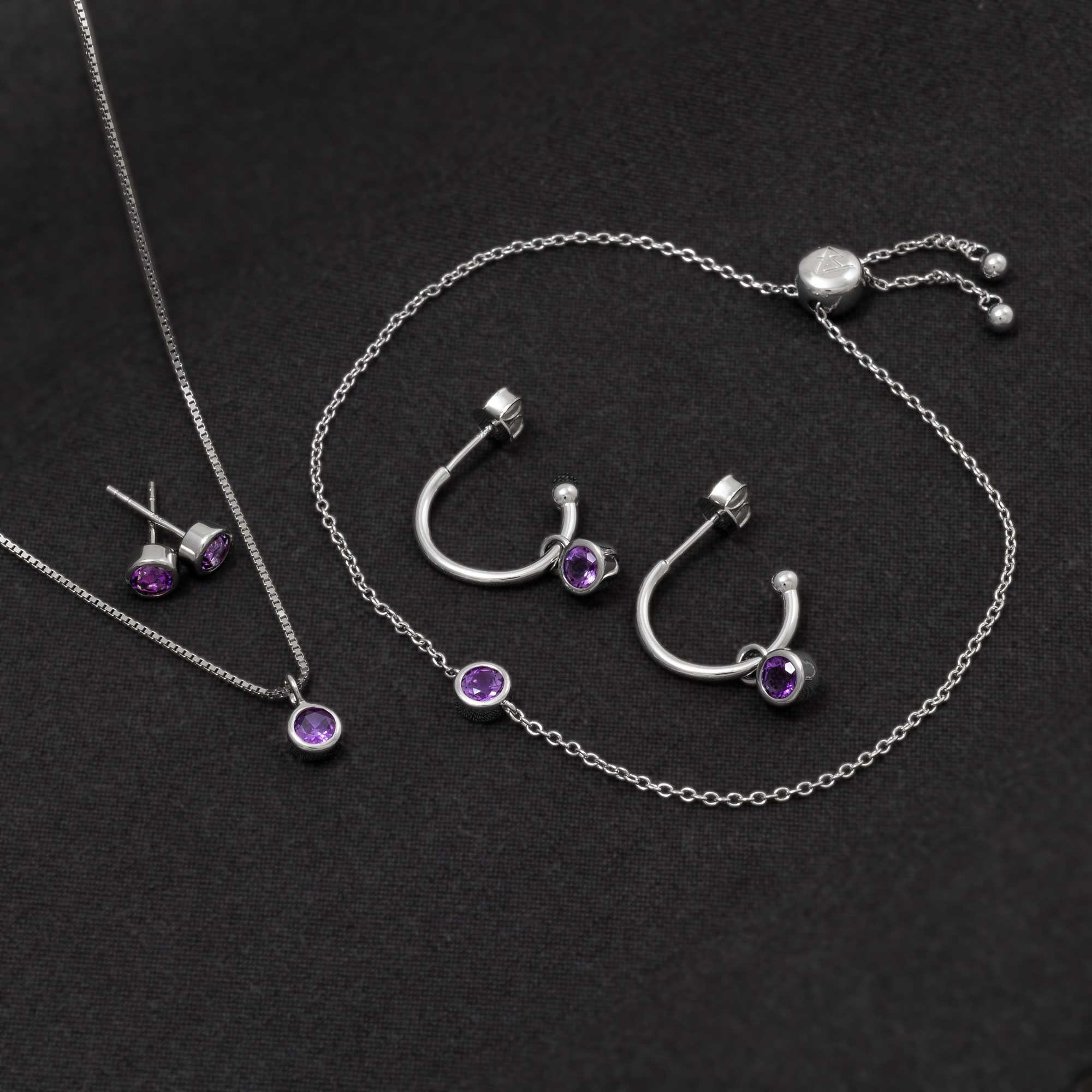Set of sterling silver jewellery featuring purple amethyst gemstones, including stud earrings, hoop earrings, a pendant necklace, and a delicate bracelet, styled on black fabric.