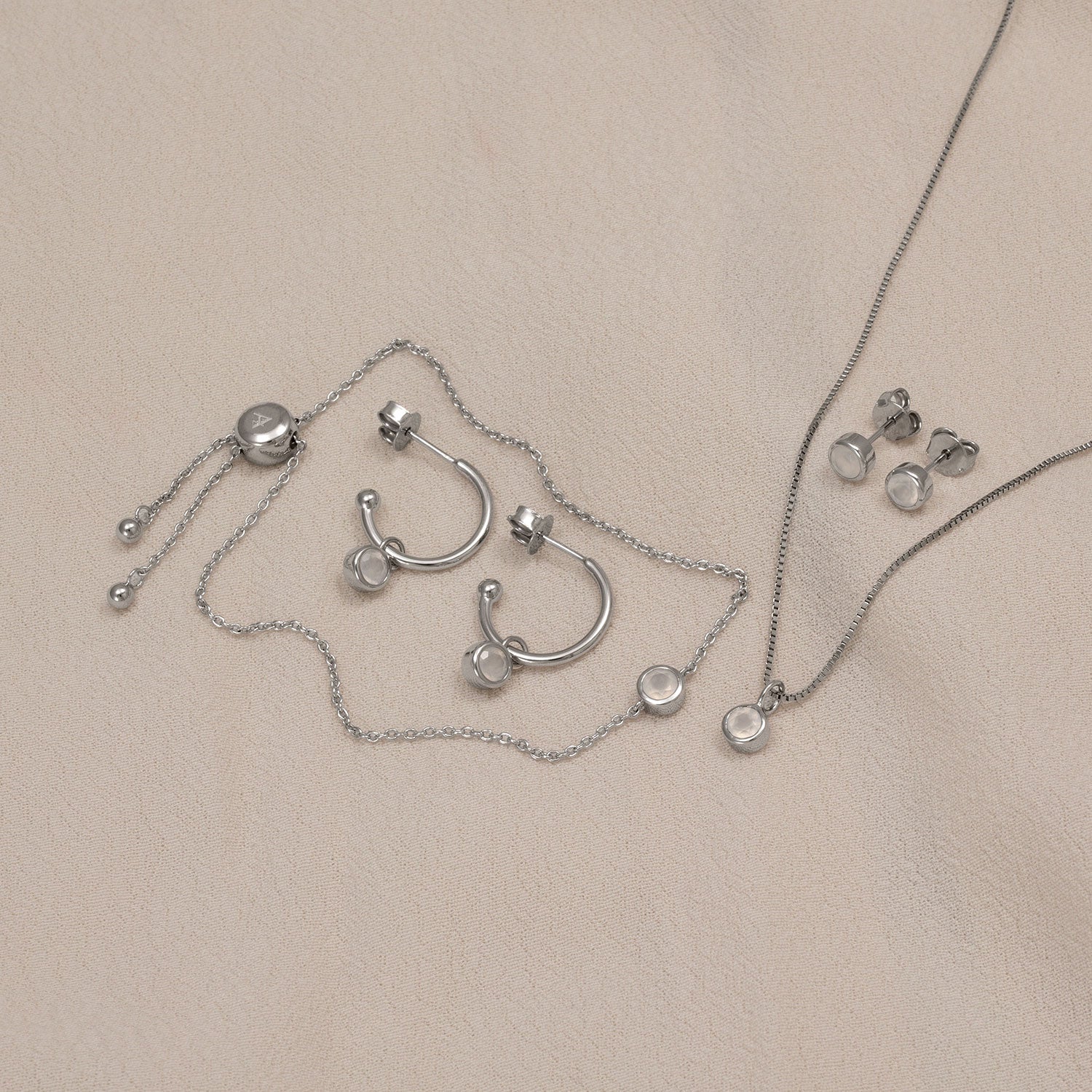 Sterling silver jewellery set featuring polished hoop earrings, delicate pendant necklace, and stud earrings each adorned with smooth aqua chalcedony stones, styled on a neutral fabric.