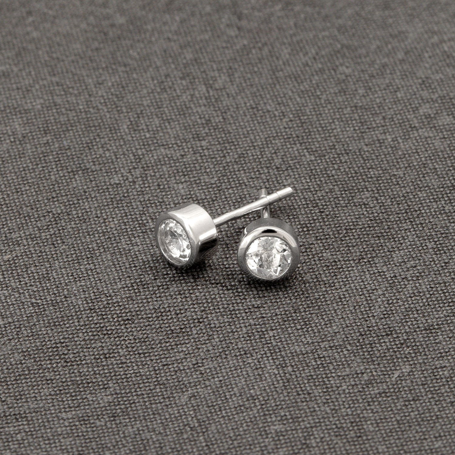 Pair of polished sterling silver stud earrings featuring bezel-set white topaz gemstones, presented on a textured grey fabric background, exemplifying refined British craftsmanship.